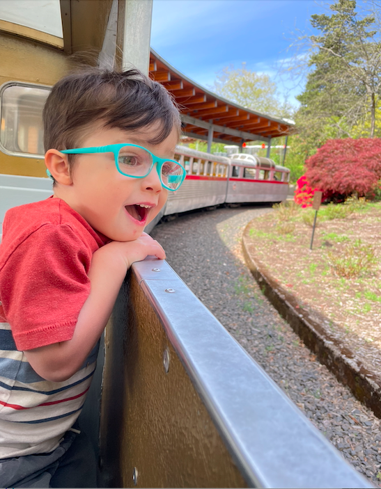 Child with glasses leaning over the side of a train, looking excited as the train rides along a track through a park with trees and a building nearby.