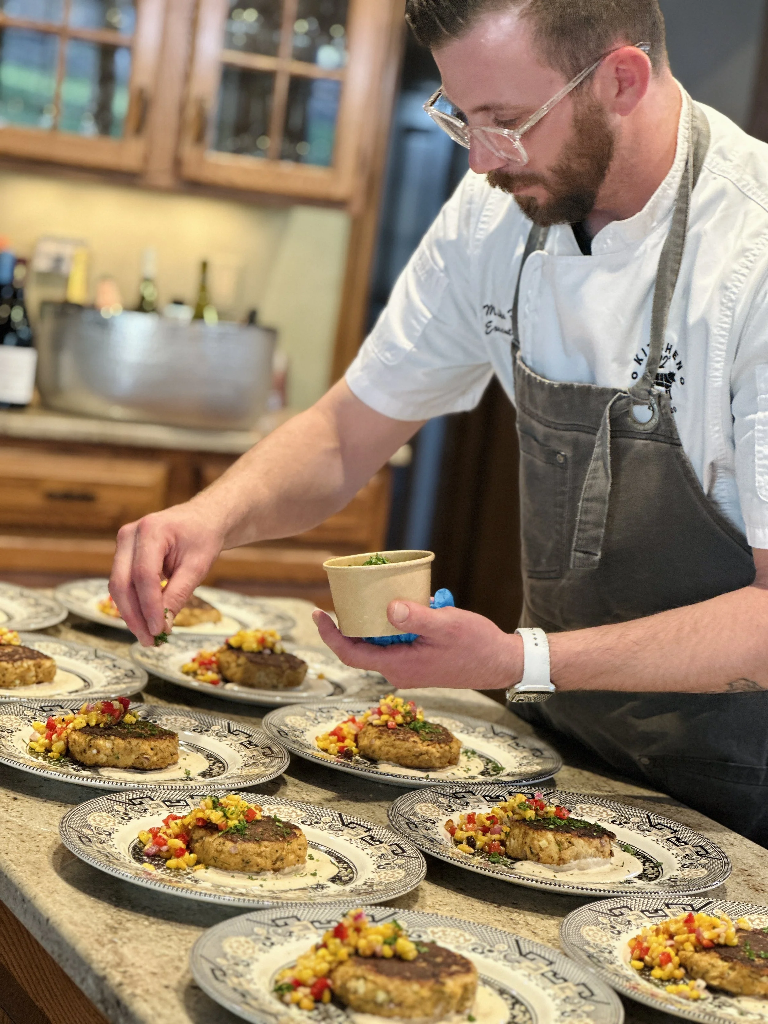 A chef garnishes plates of food with a colorful corn relish or salsa in a kitchen setting.