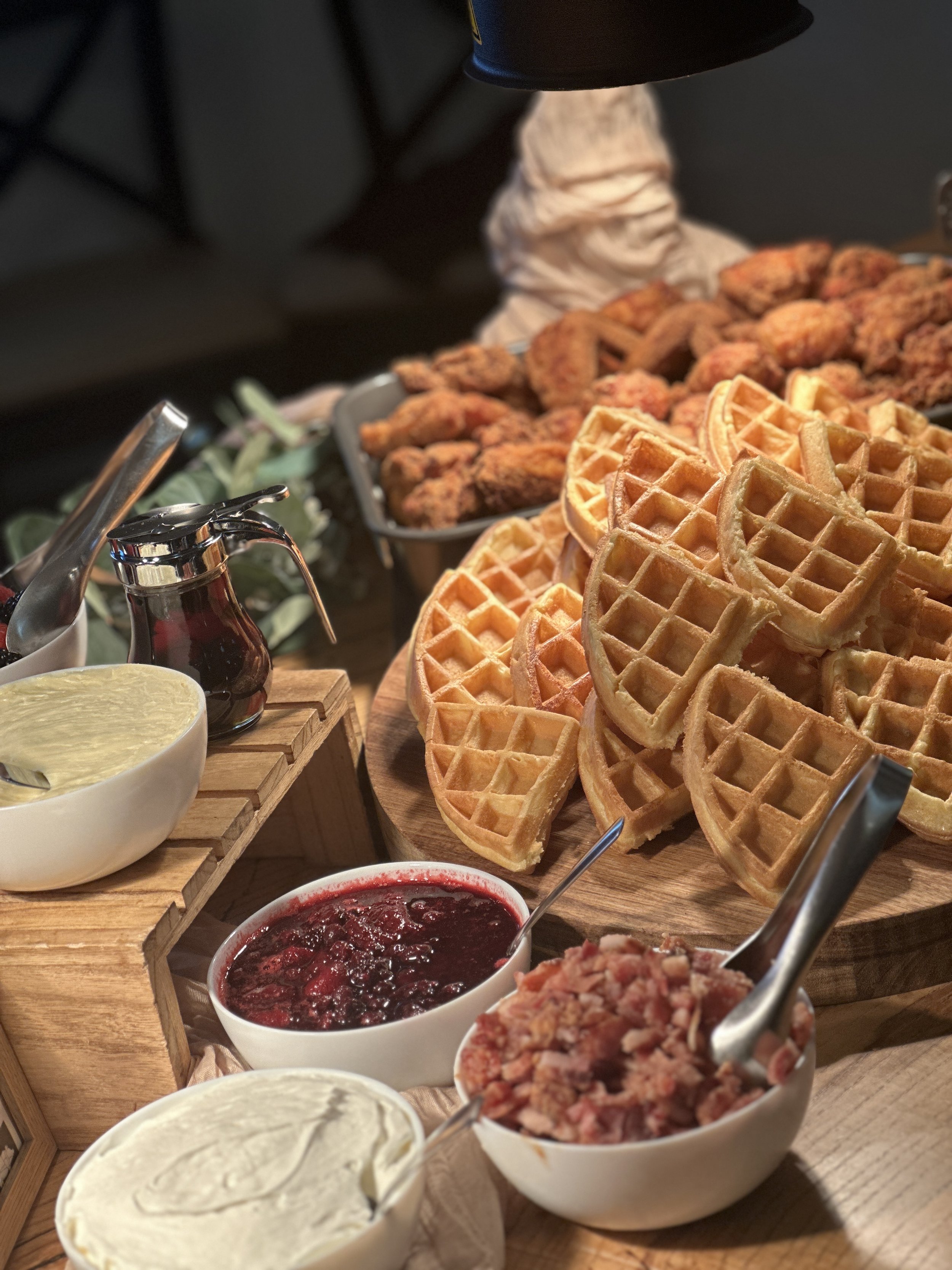 Stack of waffles with syrup, bowls of whipped cream, berry compote, and bacon on a wooden platter.
