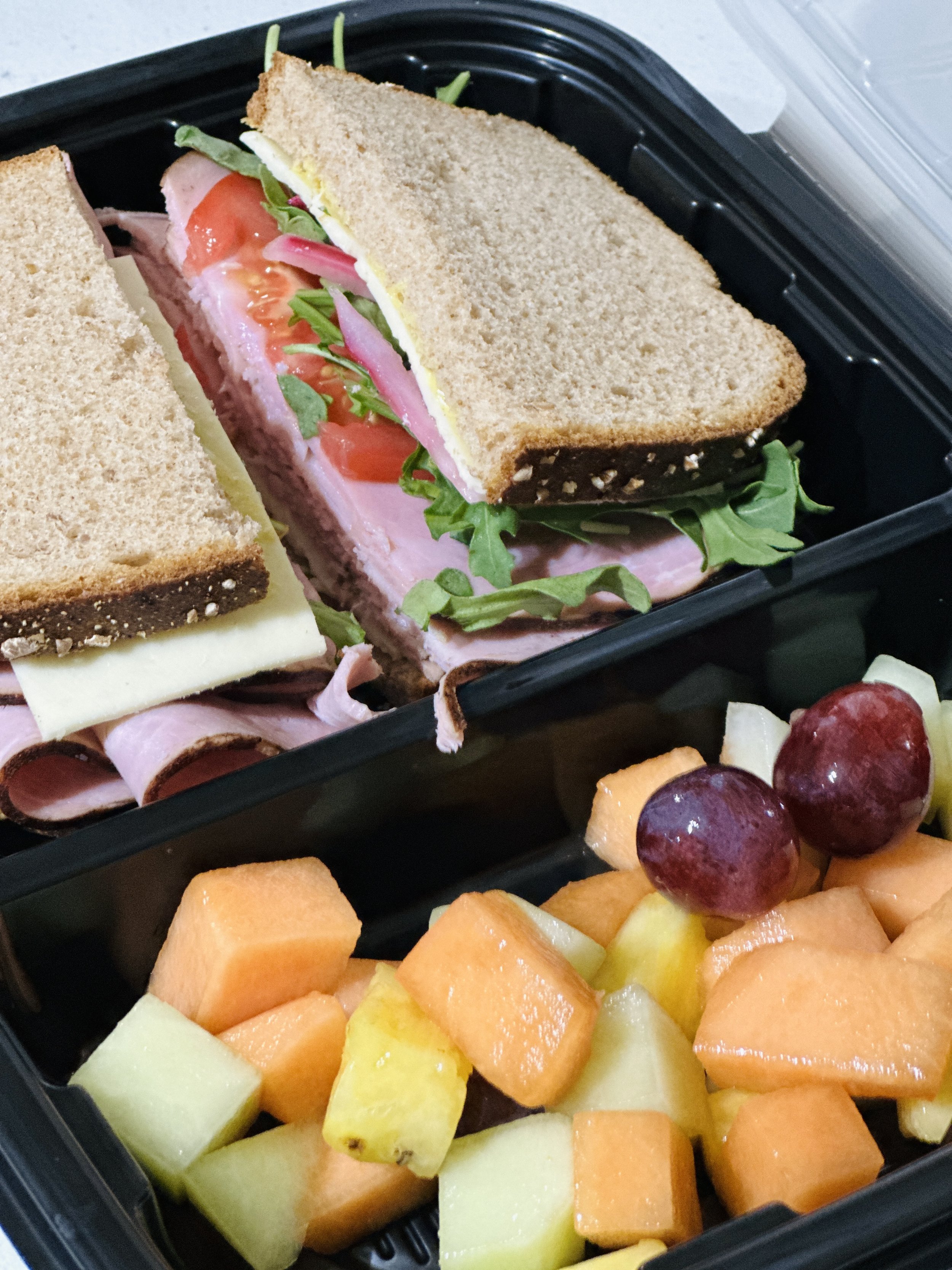 A black lunchbox containing a sandwich with ham, tomato, and arugula, and a side of chopped cantaloupe, honeydew, and red grapes.