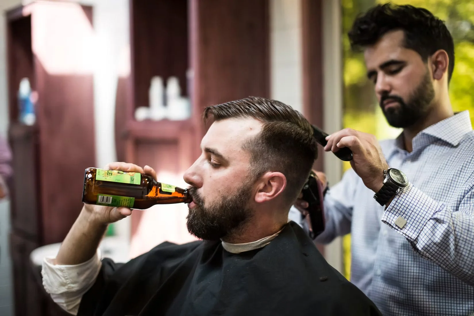 A groom drinks a beer while a Topshelf Grooming barber styles his hair before the wedding ceremony.