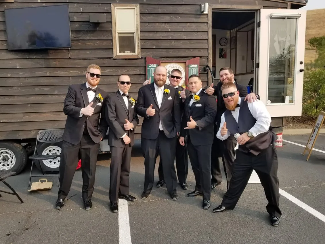 A groom and his groomsmen in tuxedos pose outside the Topshelf Grooming mobile barbershop trailer after their pre-wedding haircuts.