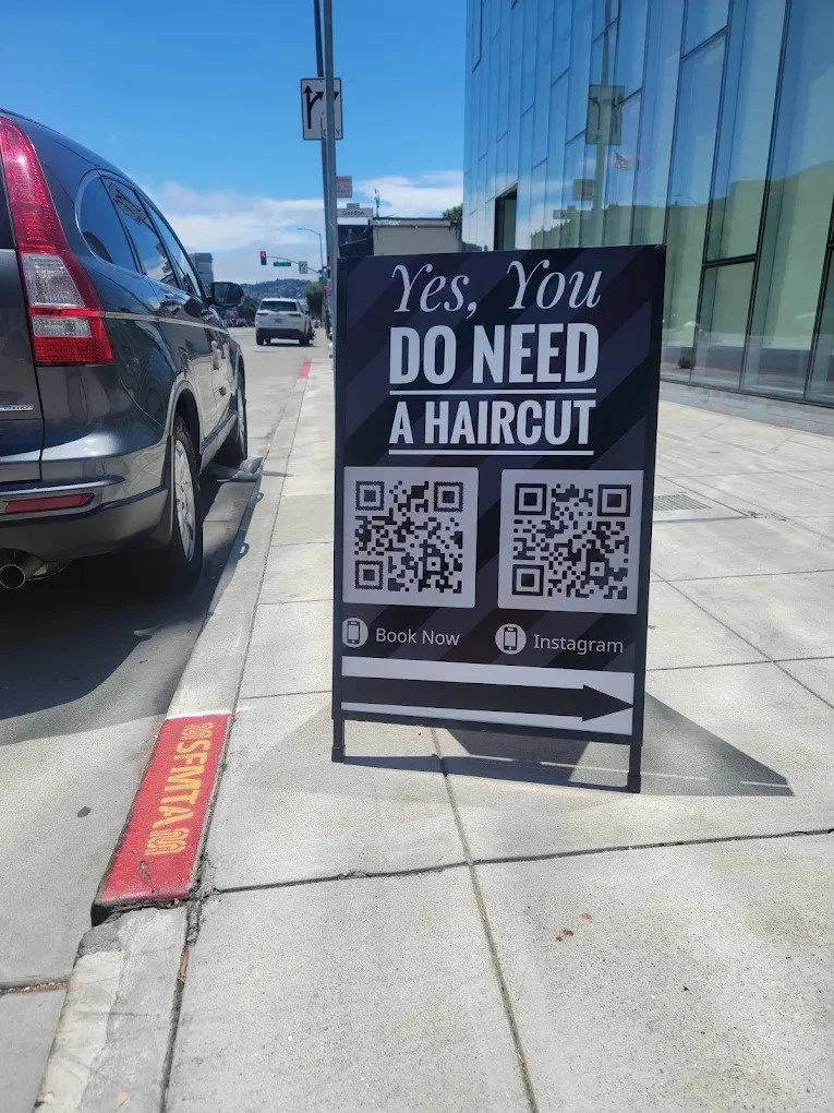 The Bus Stop Barbershop's a-frame sign on the Harrison Street sidewalk in SoMa, San Francisco, reading "Yes, You Do Need a Haircut."