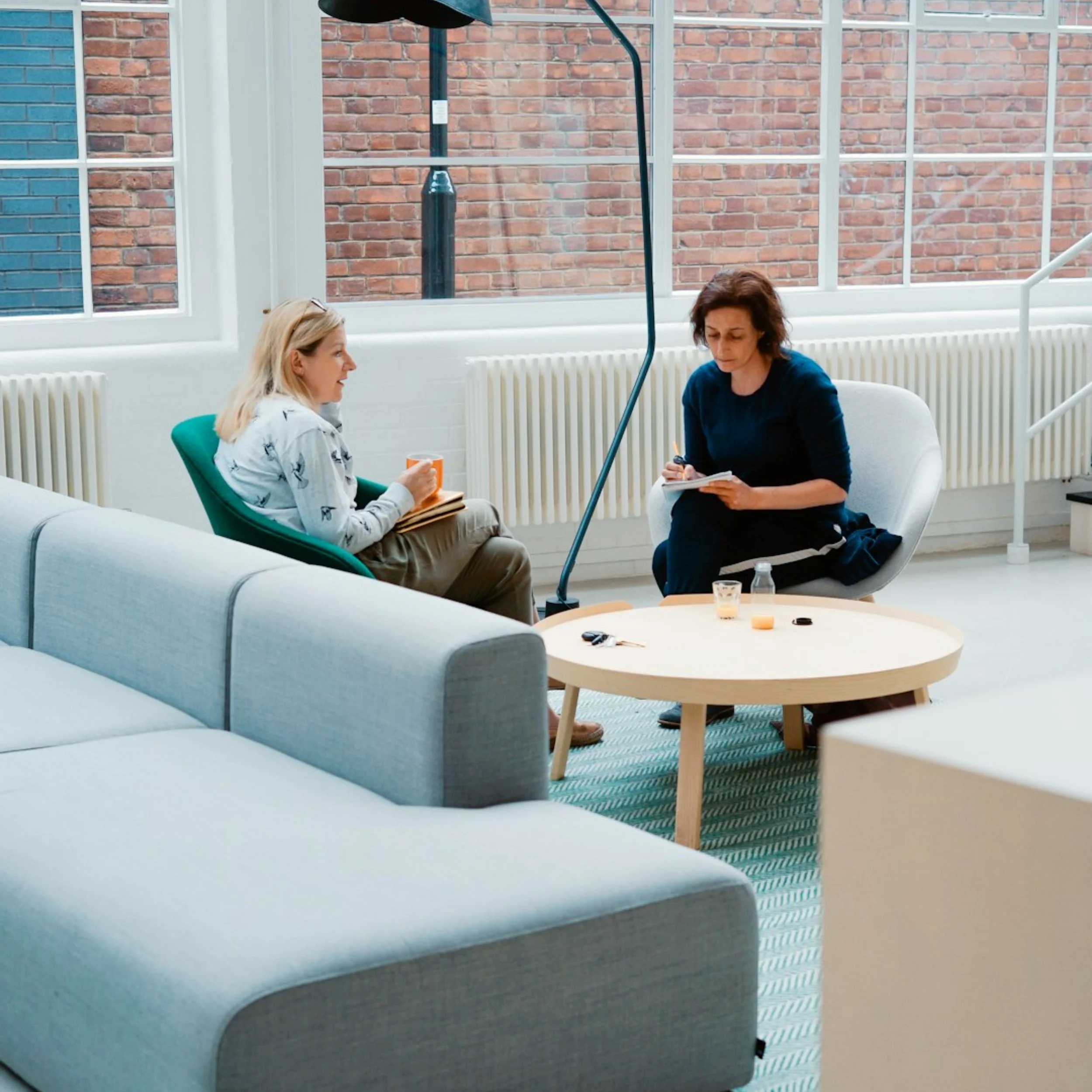 Two women are seated on a couch having a conversation in a brightly lit room with large windows and brick walls. One woman is blonde, wearing a light jacket, and holding a notebook, while the other has dark hair, dressed in a dark top, and is taking notes.