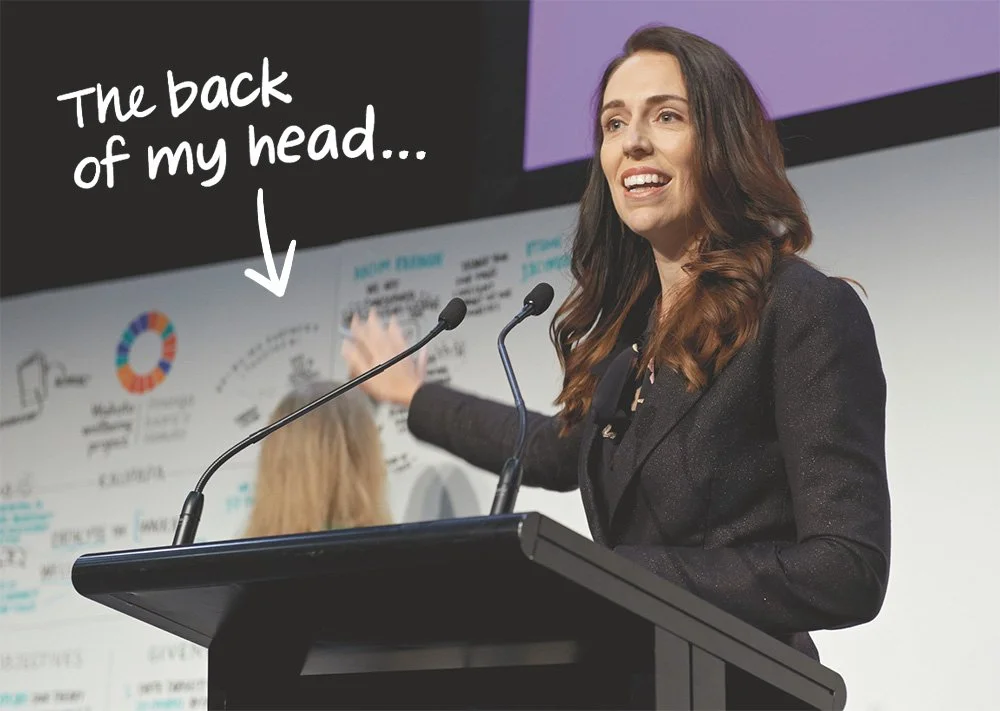 A national leader is giving a presentation, and gesturing to the large whiteboard wall behind her where Lisa is working with her back to the audience on a large graphic recording with handwritten notes and drawings.