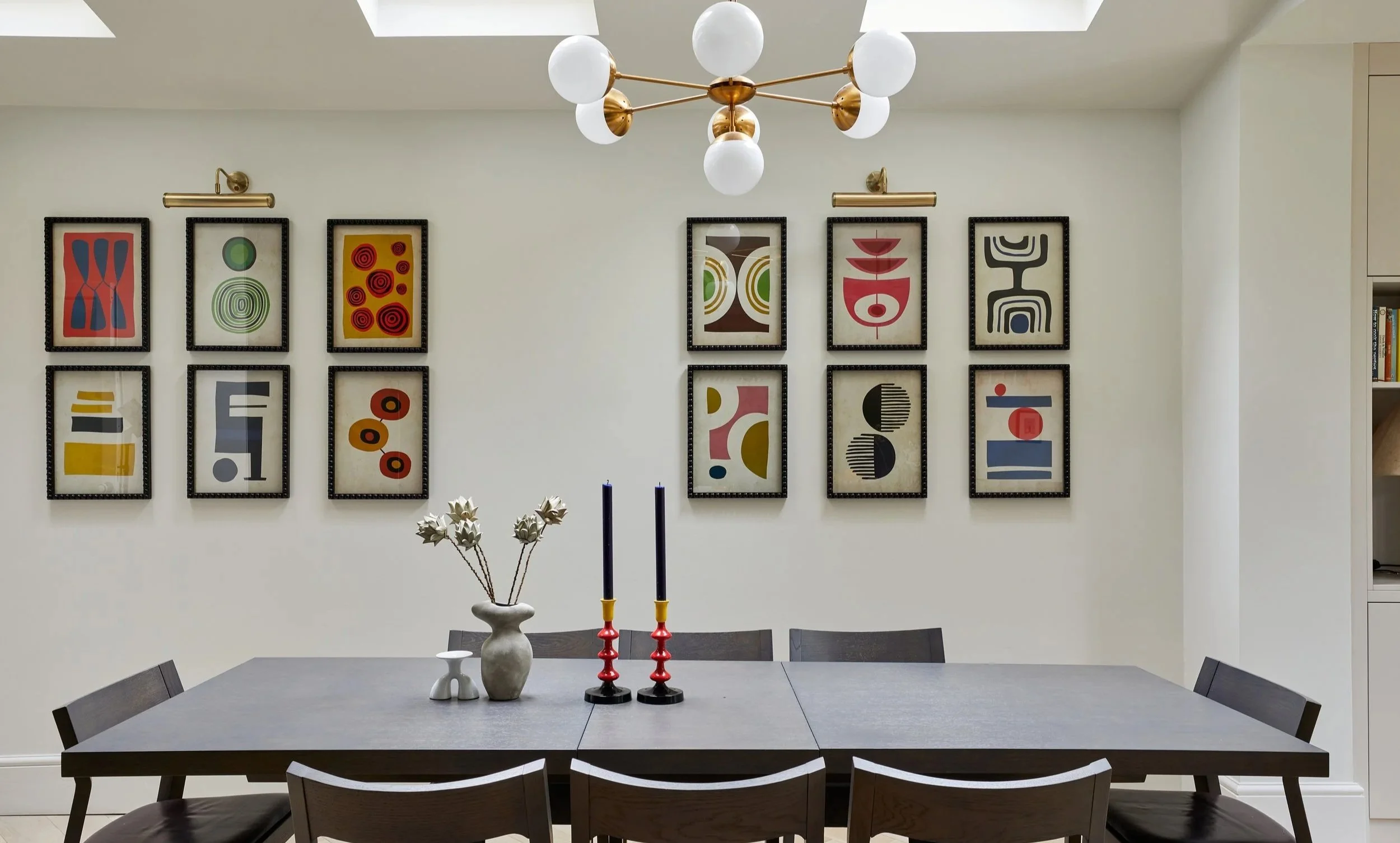 A modern dining room with a dark wooden table and chairs. On the table, there are three decorative vases and two red candlesticks. The wall features a series of colorful abstract framed art pieces evenly spaced. A contemporary chandelier with white g