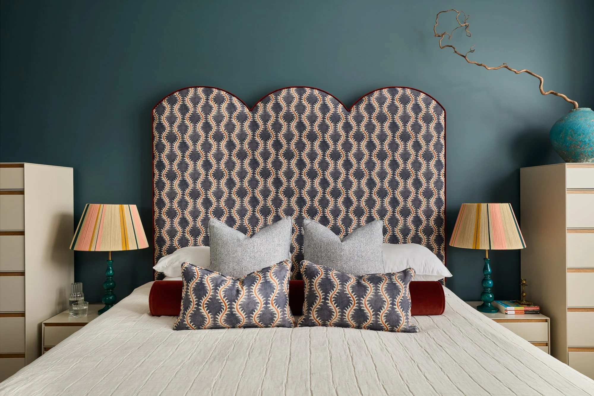 Bespoke-designed headboard in a contemporary patterned fabric with matching scatter cushions, taupe and oak bedside furniture, and matching table lamps with turquoise ceramic bases and colourful striped shades in a Chiswick master bedroom.