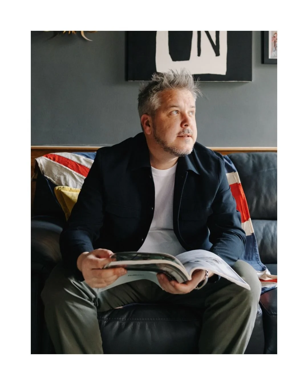 A man with gray hair and a beard sitting on a black leather couch, holding a magazine, with a neutral expression, wearing a black jacket and gray pants, in a room with dark gray walls and framed artwork.