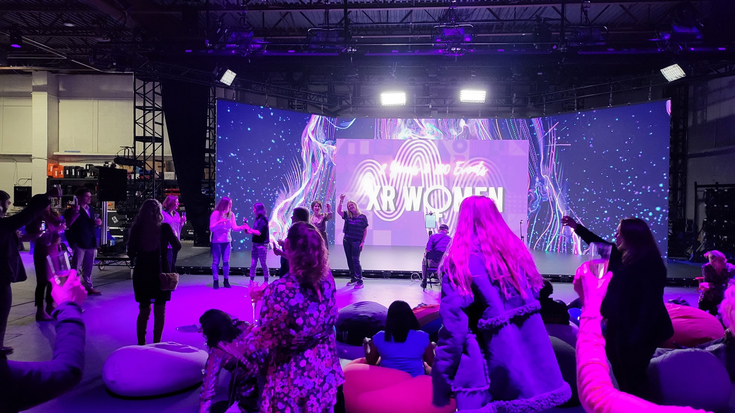 People gathered at an event with a digital backdrop displaying 'XR Women', illuminated by purple lighting.