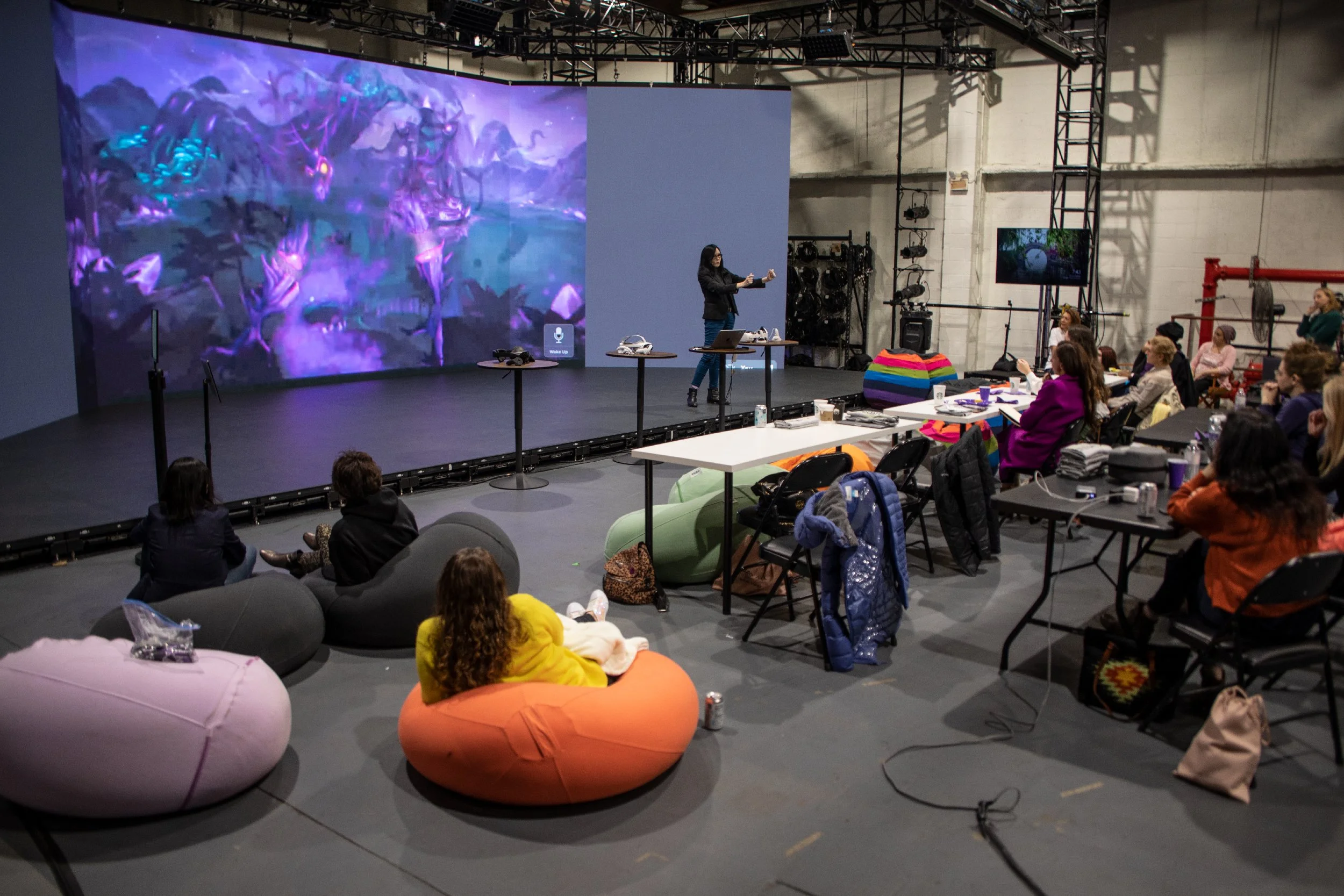 A presentation in a studio with people seated on bean bags and chairs. A large screen displays a colorful, abstract landscape. A speaker stands near the screen, addressing the audience.