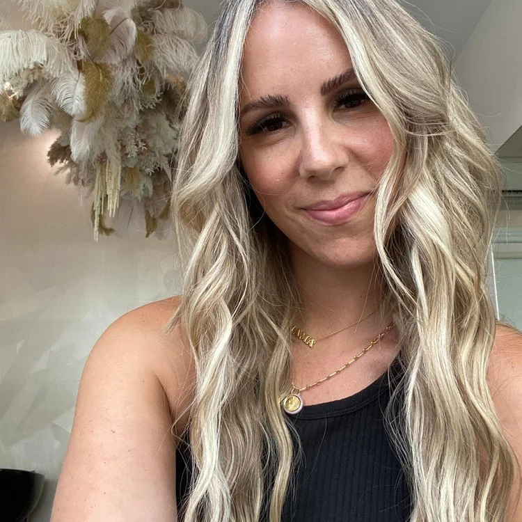 Close-up of a woman with long, wavy blonde hair smiling at the camera, wearing a black sleeveless top, gold jewelry, and standing in front of a wall with a decorative feather arrangement.