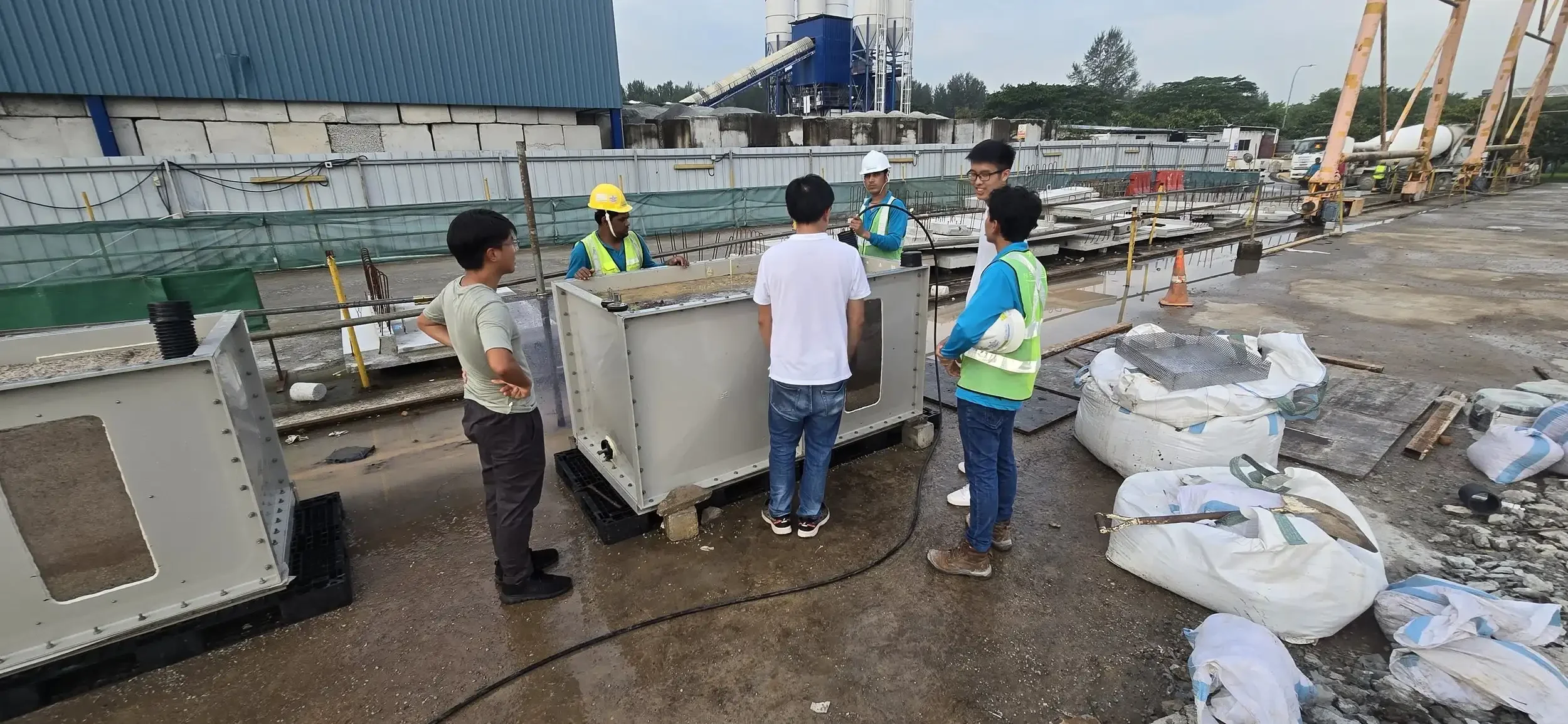 Group of construction workers and engineers at a construction site discussing a large metallic container or equipment. Some are wearing helmets and safety vests, while others are in casual clothing. The construction area has various materials, tools, and machinery, with some with large bags of supplies and a partly constructed structure in the background.