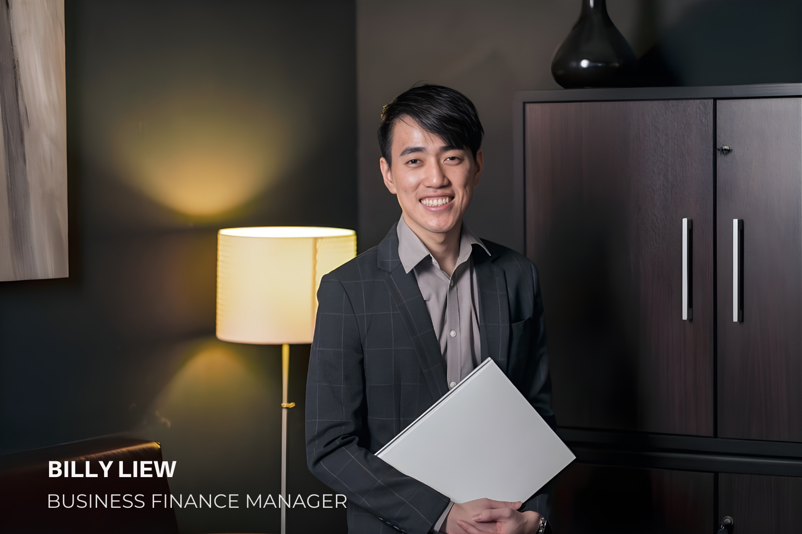 A young man with black hair, wearing a dark checkered blazer and gray shirt, smiling and holding a gray folder in a modern office or conference room. Behind him is a dark wooden cabinet and a table lamp with a yellow shade, illuminating a dark-colore