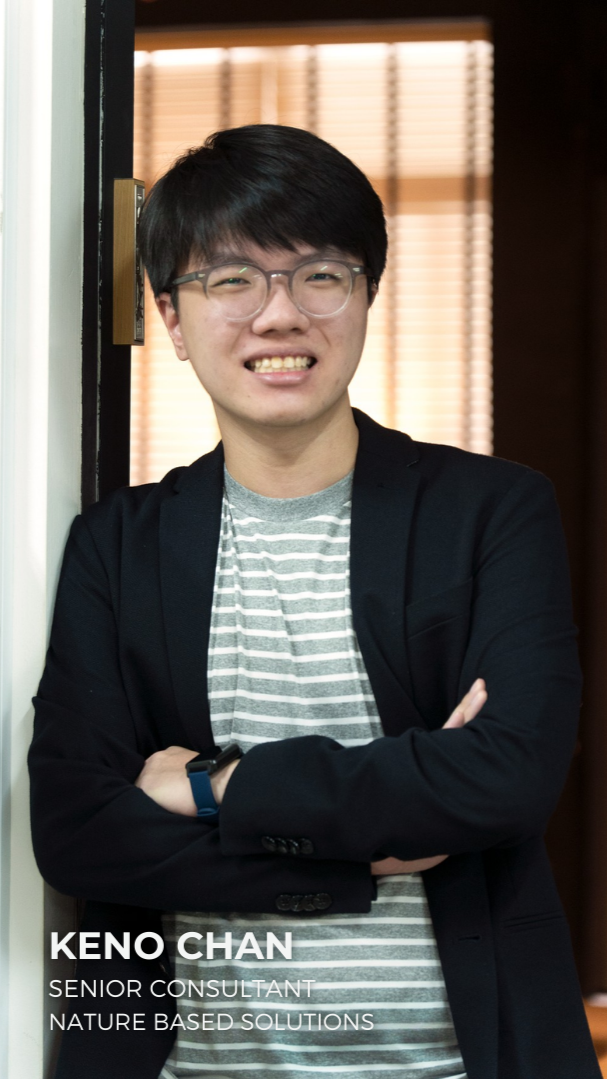 A young man wearing glasses, a striped t-shirt, and a black blazer, standing with arms crossed in front of a doorframe with wooden blinds in the background, smiling at the camera. Text overlay: Keno Chan, Senior Consultant, Nature Based Solutions.