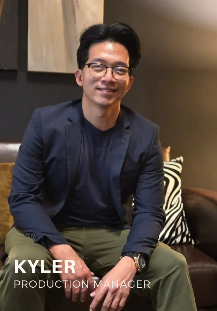 A young man with black hair and glasses, wearing a black blazer, black t-shirt, and khaki pants, sitting on a brown leather couch in a dimly lit room with abstract artwork on the wall behind him. The text overlay identifies him as Kyler, a Production
