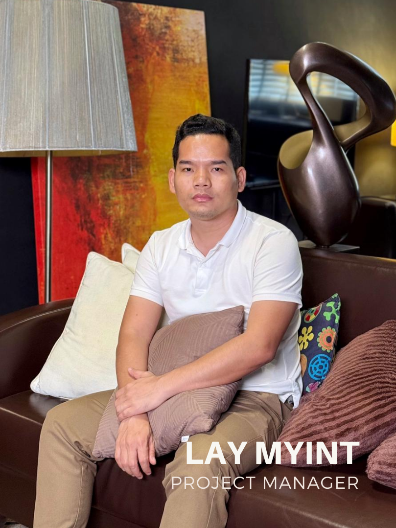 A man wearing a white polo shirt sitting with arms crossed in an office setting. Text overlay reads "Lay Myint, Project Manager."