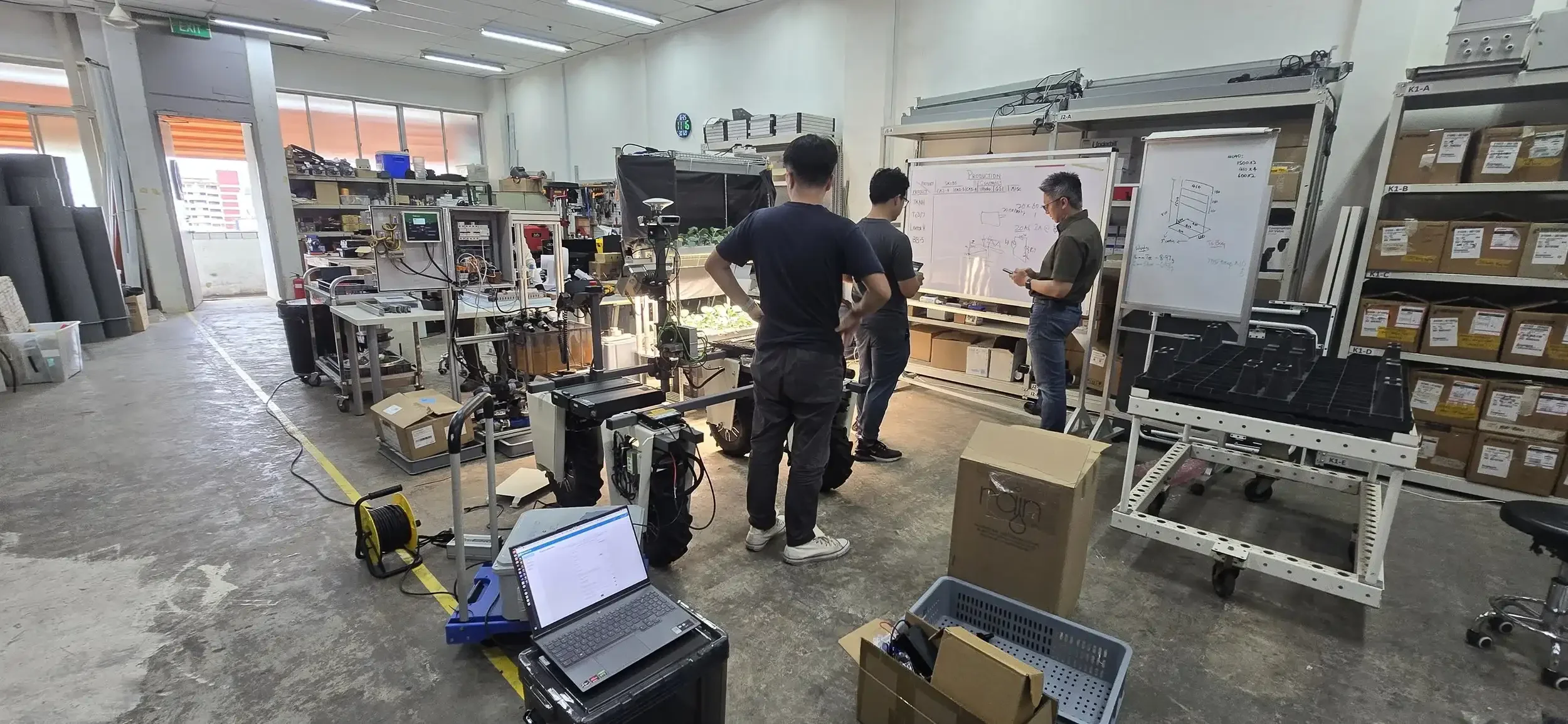 Three people working in a machine shop with various equipment, tools, and storage shelves around them, some whiteboards with sketches and notes, and a laptop on a nearby table.