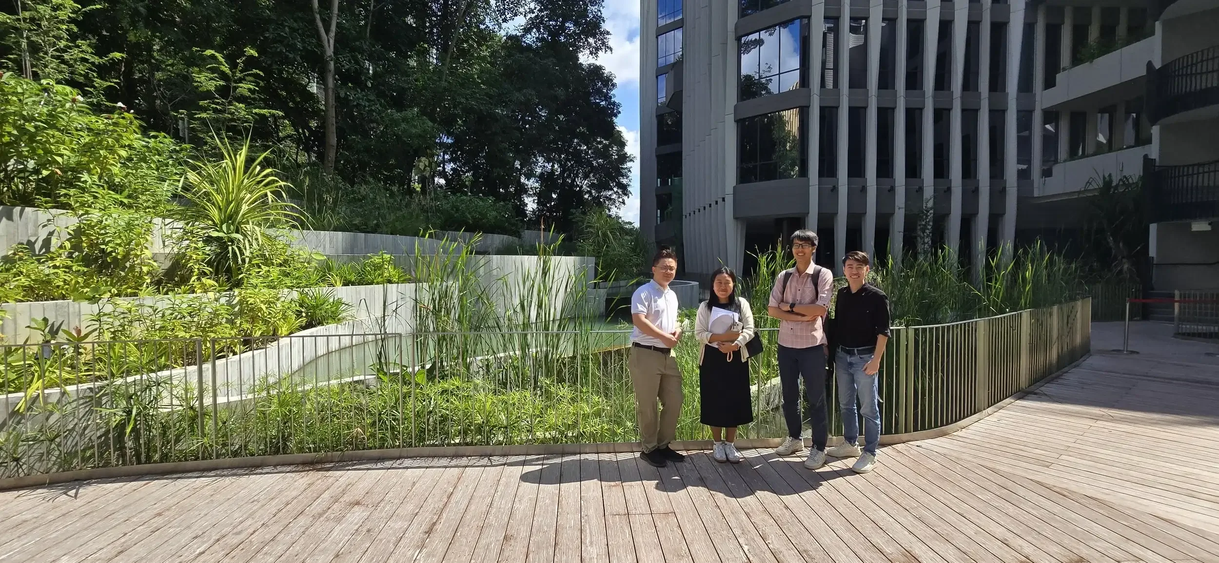 Four young adults standing on a wooden pathway in front of lush greenery, with a modern building with tall glass windows in the background.