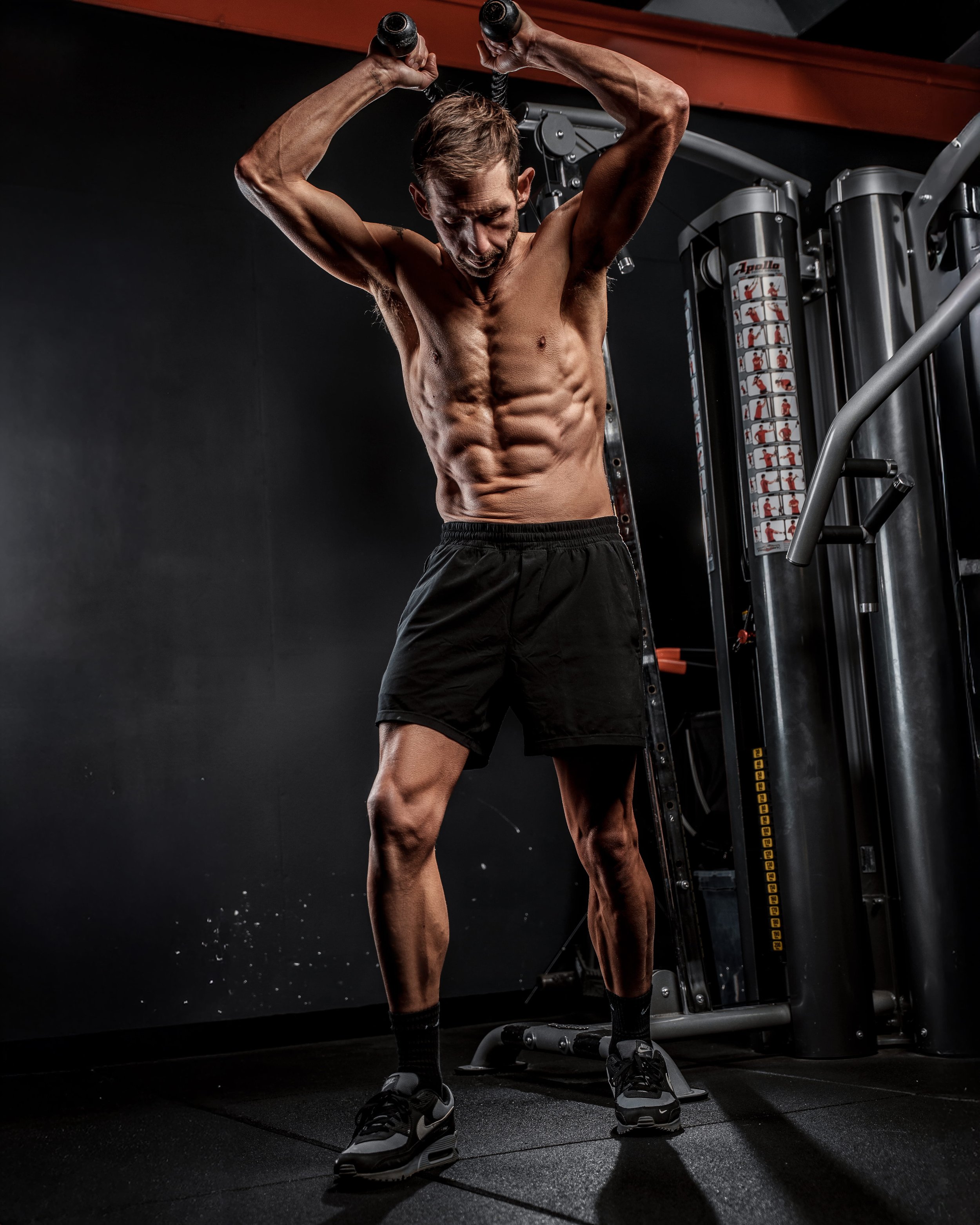 A man with a muscular build doing an overhead triceps extension exercise at the gym, shirtless and wearing black shorts and sneakers.