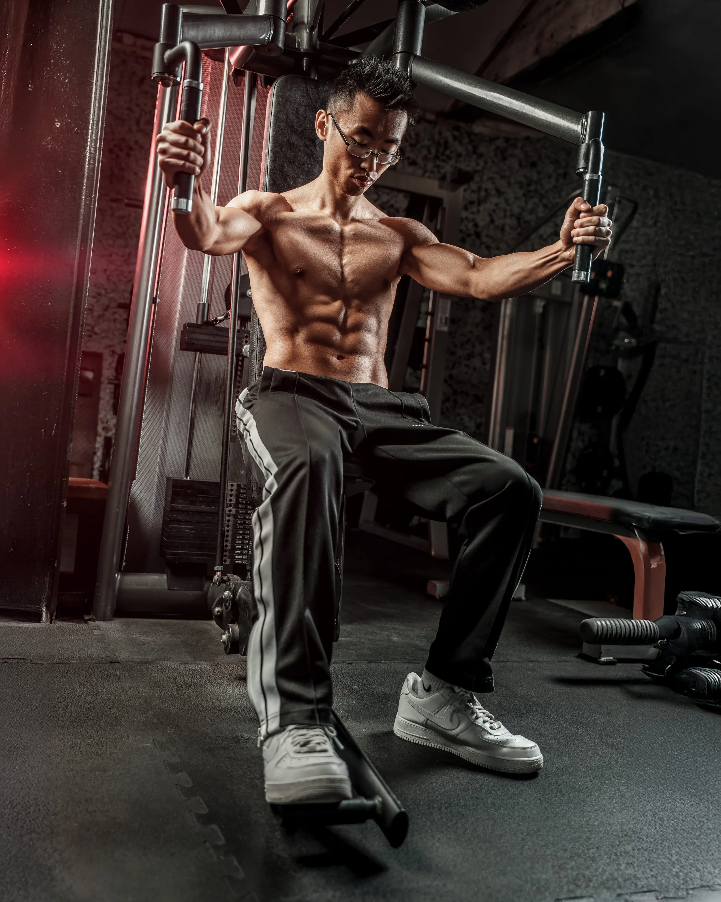 Bodybuilding photoshoot of a shirtless male athlete using a cable machine in a dark private gym, emphasizing upper body muscle definition