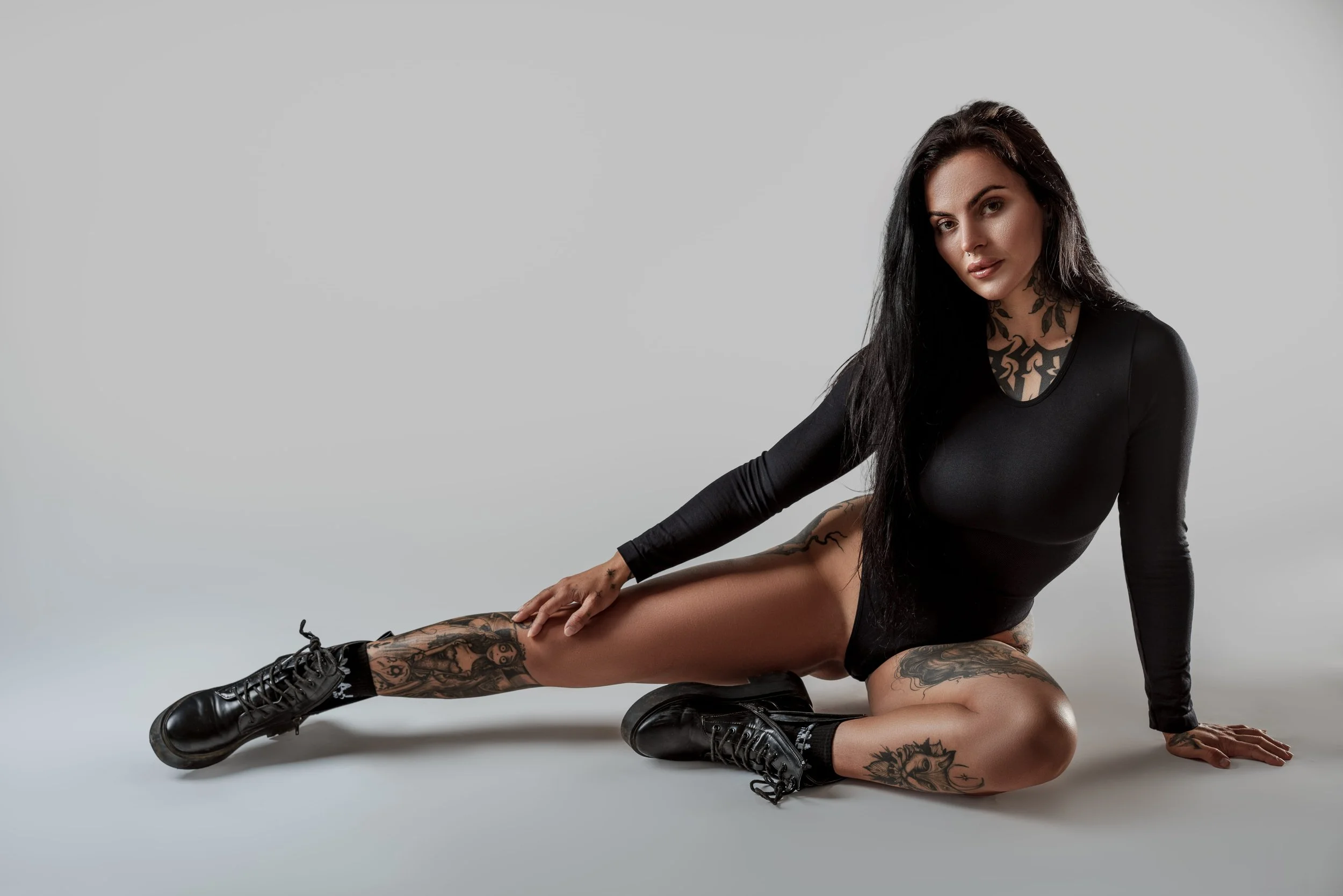 Editorial boudoir photography studio portrait of a tattooed woman seated on the floor with an inverted leg pose showing strength posture and calm confident presence