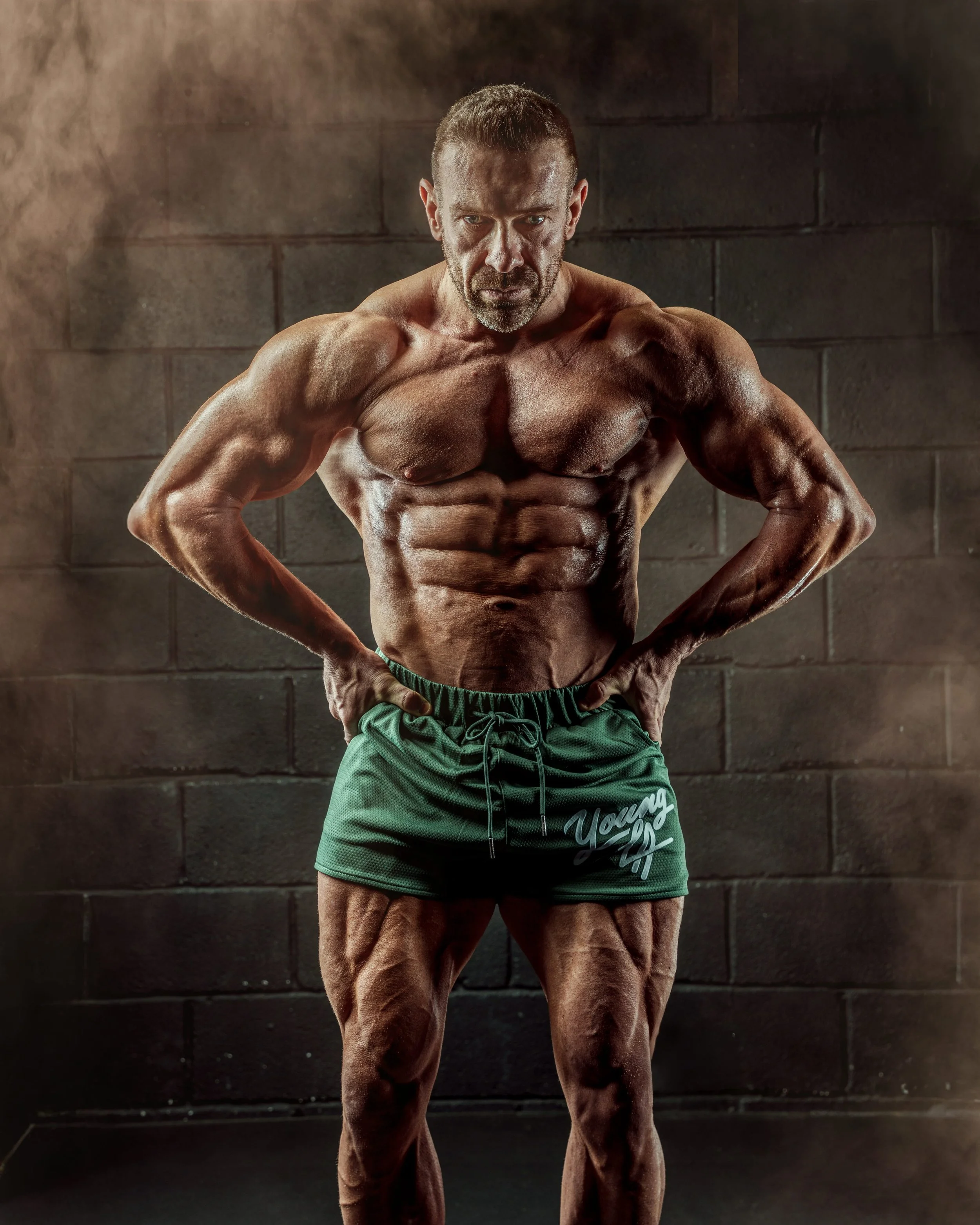 A muscular man posing indoors with hands on hips, wearing green shorts, in front of a dark brick wall.
