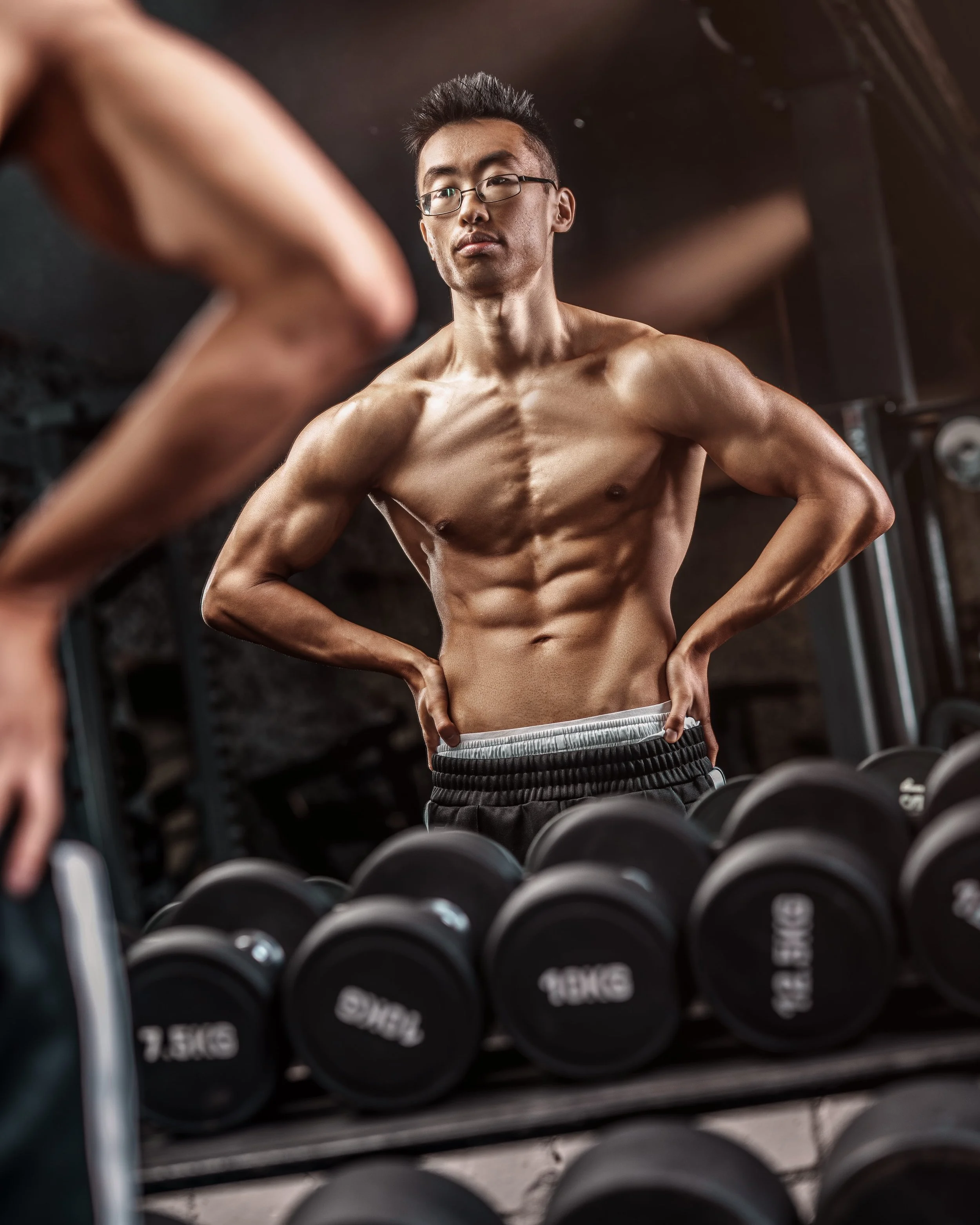 A shirtless young man with glasses at a gym, standing in front of a mirror with his hands on his hips, surrounded by dumbbells.