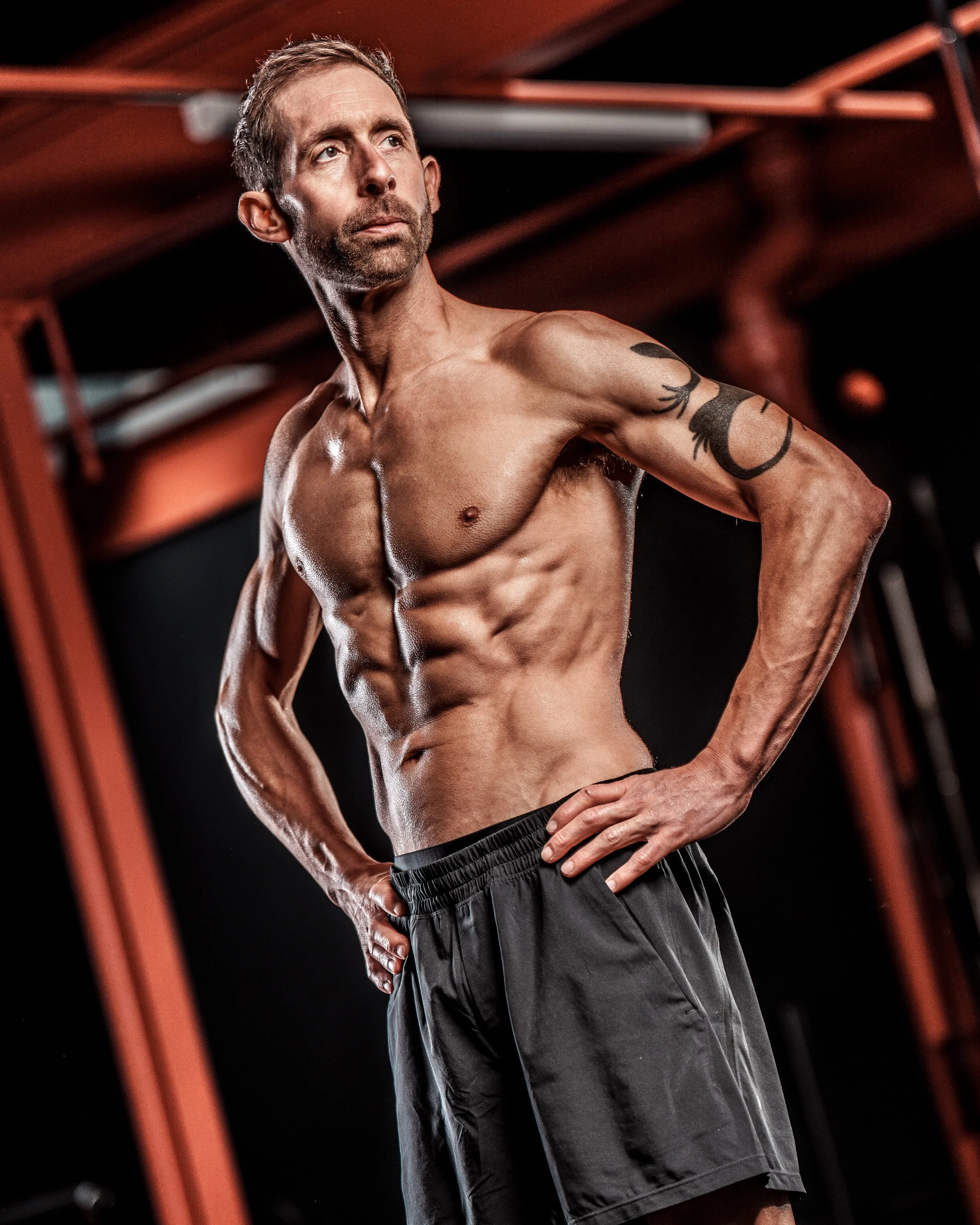 A shirtless man with a muscular physique, tattoos on his right arm, standing in a gym environment with his hands on his hips.