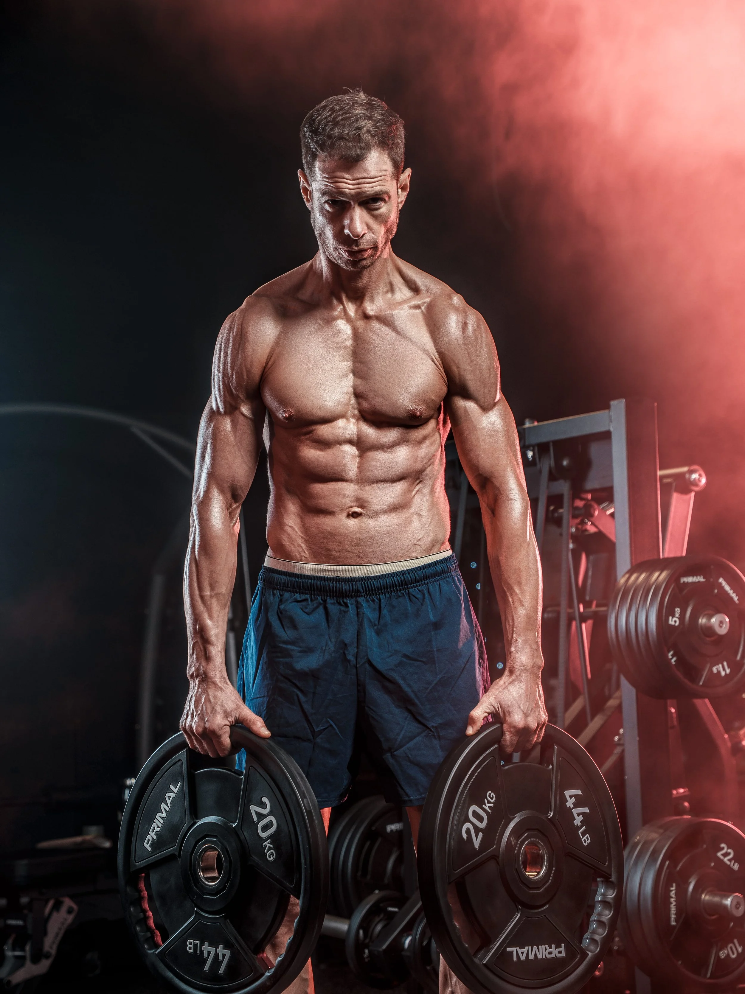 A muscular man with defined abs lifting weight plates in a gym, with weights and gym equipment visible in the background.