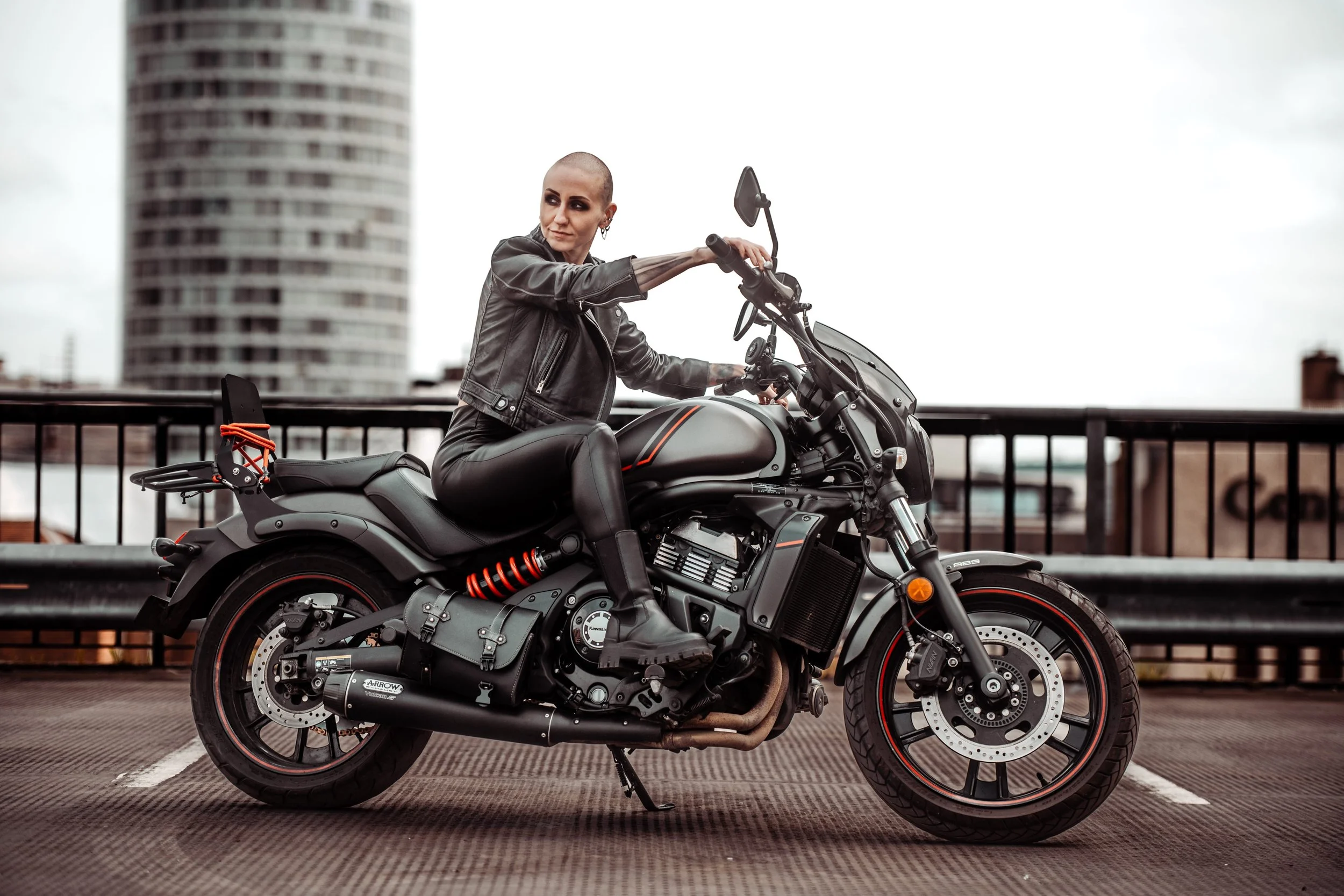 A woman with a shaved head wearing a black leather jacket and black pants sits on a black motorcycle on a rooftop parking lot with a city skyline in the background.