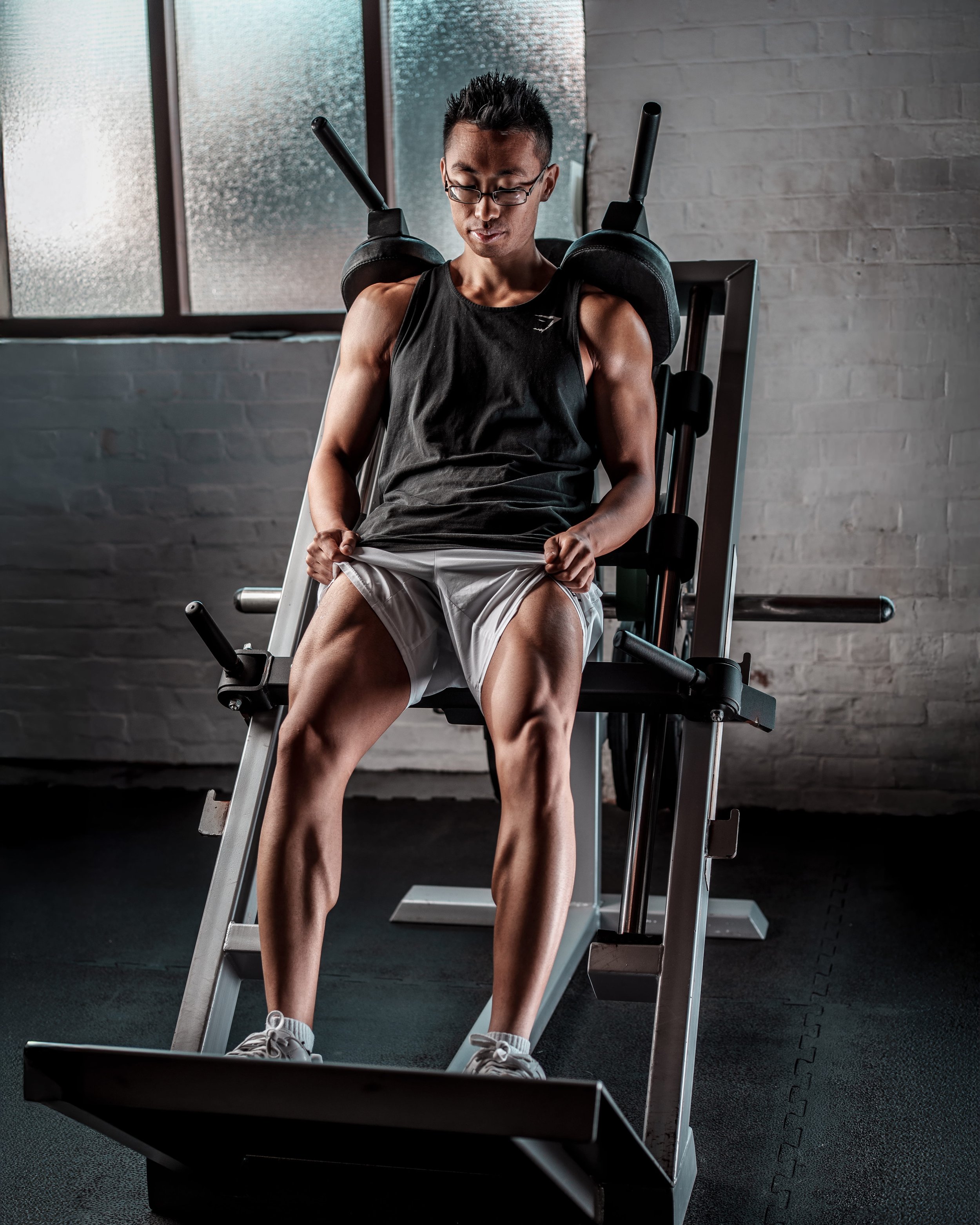 A young man with short dark hair, glasses, wearing a black tank top and white shorts, sitting on a leg press machine in a gym, lifting his shorts to reveal muscular thighs.