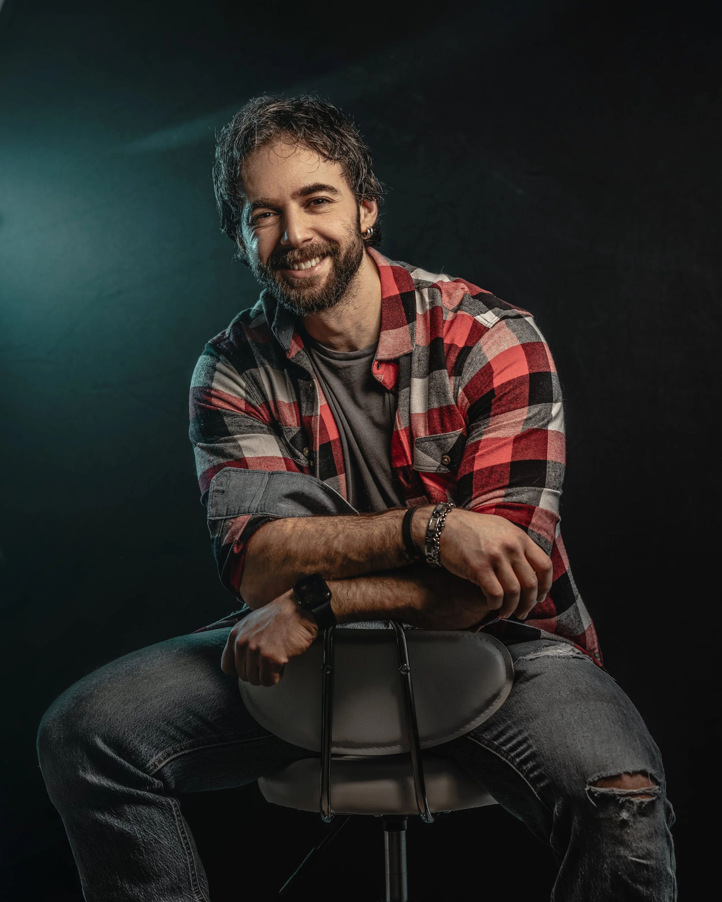 A smiling man with a beard and curly hair, wearing a red and black plaid shirt over a gray t-shirt, sitting on a stool with arms crossed, against a dark background.
