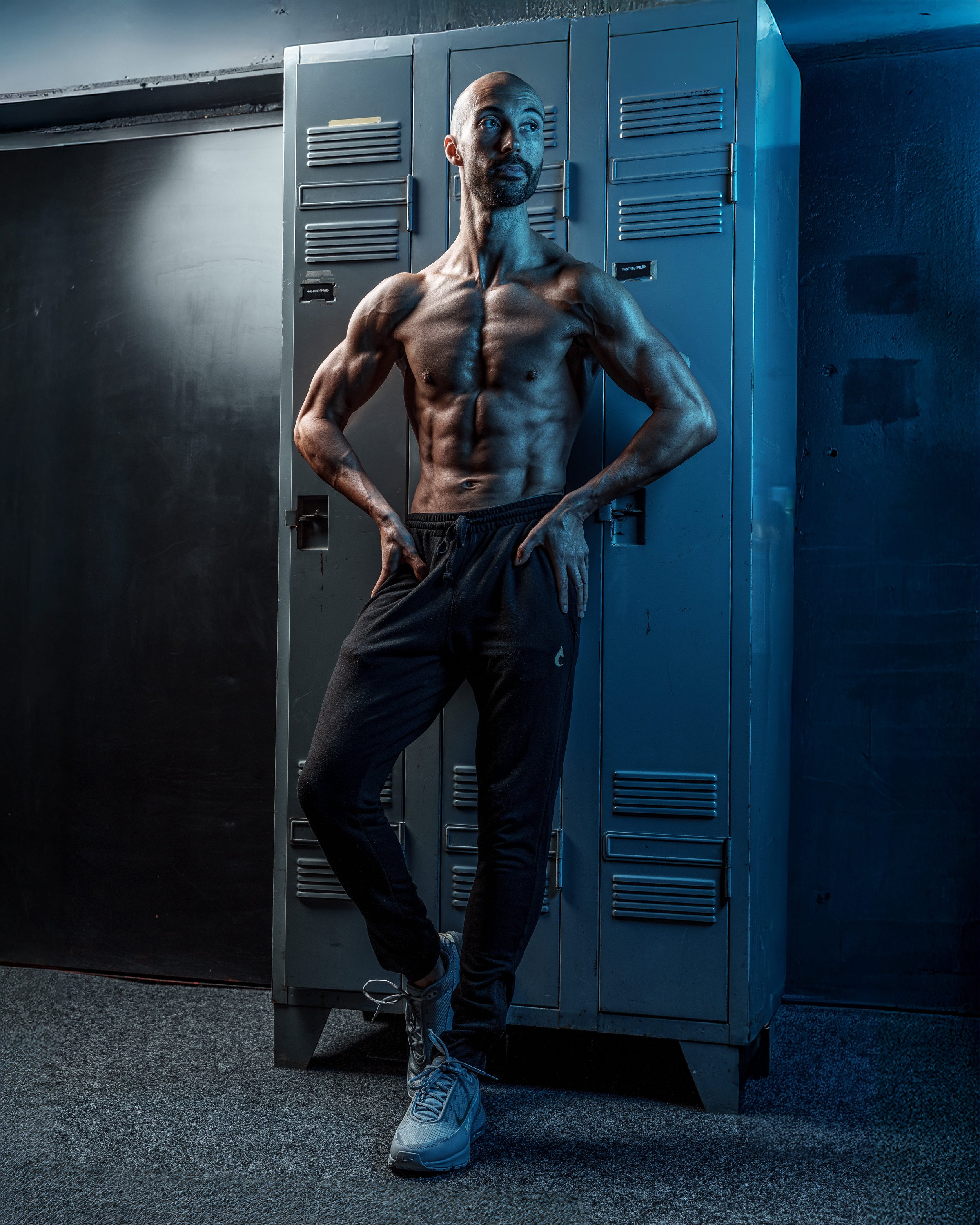 Bodybuilding photoshoot of a lean muscular man standing shirtless in a locker room, photographed with dramatic low light for physique emphasis.