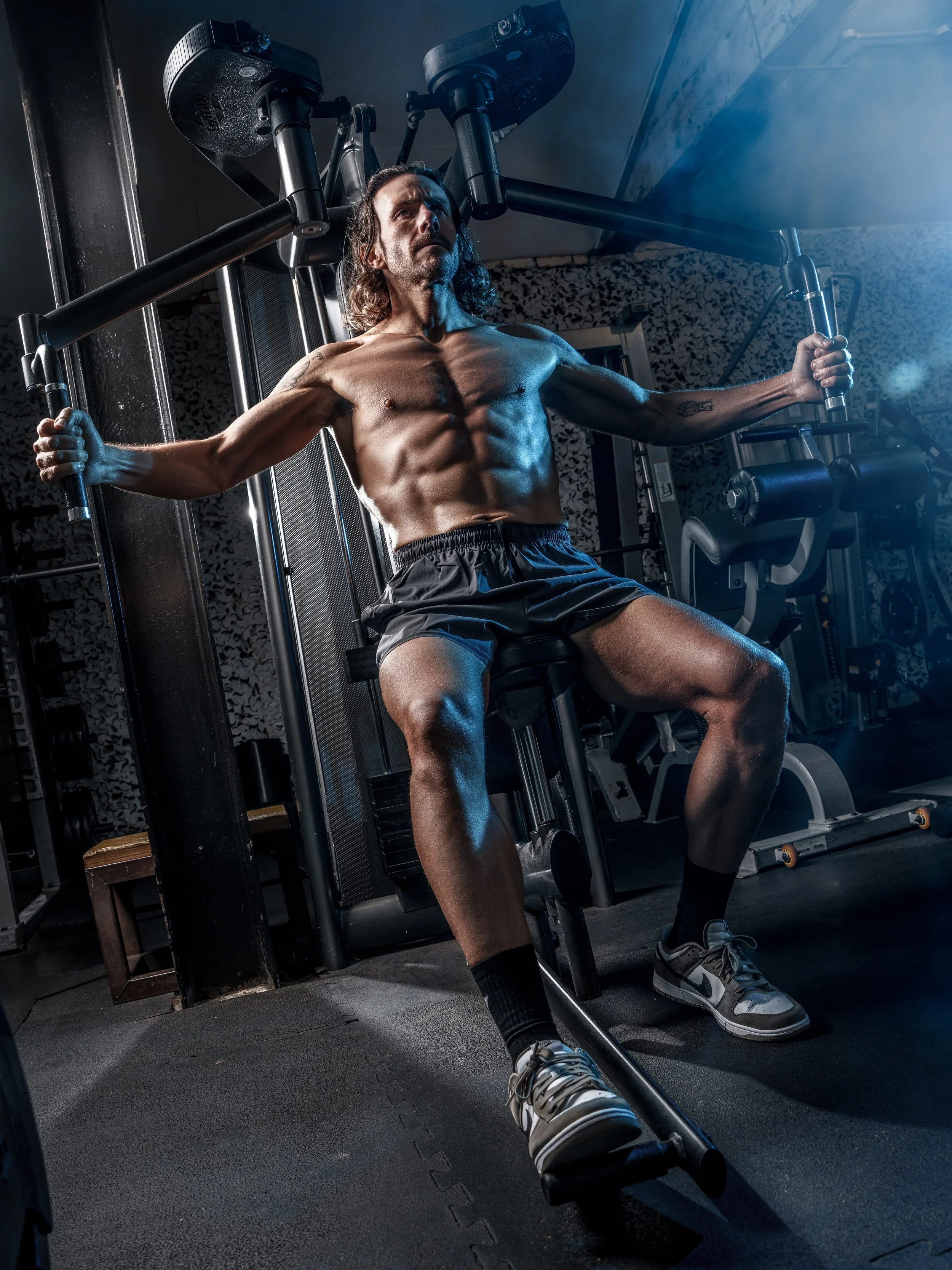 A muscular man with long hair, shirtless, wearing black shorts, sneakers, and black socks, sits on exercise equipment in a gym, holding onto handles and leaning back with a serious expression.