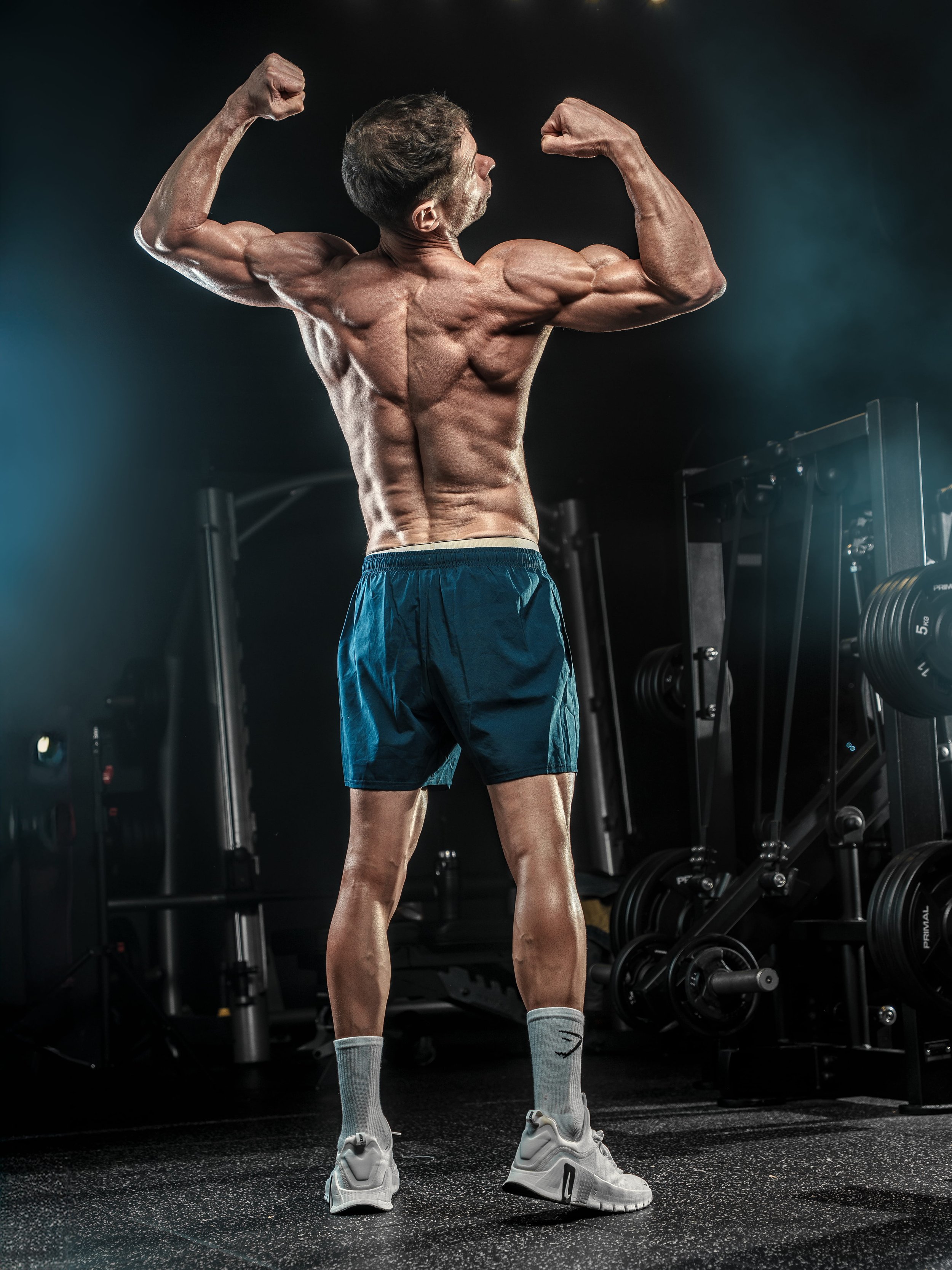 A muscular man in a gym flexing his back and arm muscles while wearing blue shorts, gray socks, and white sneakers.