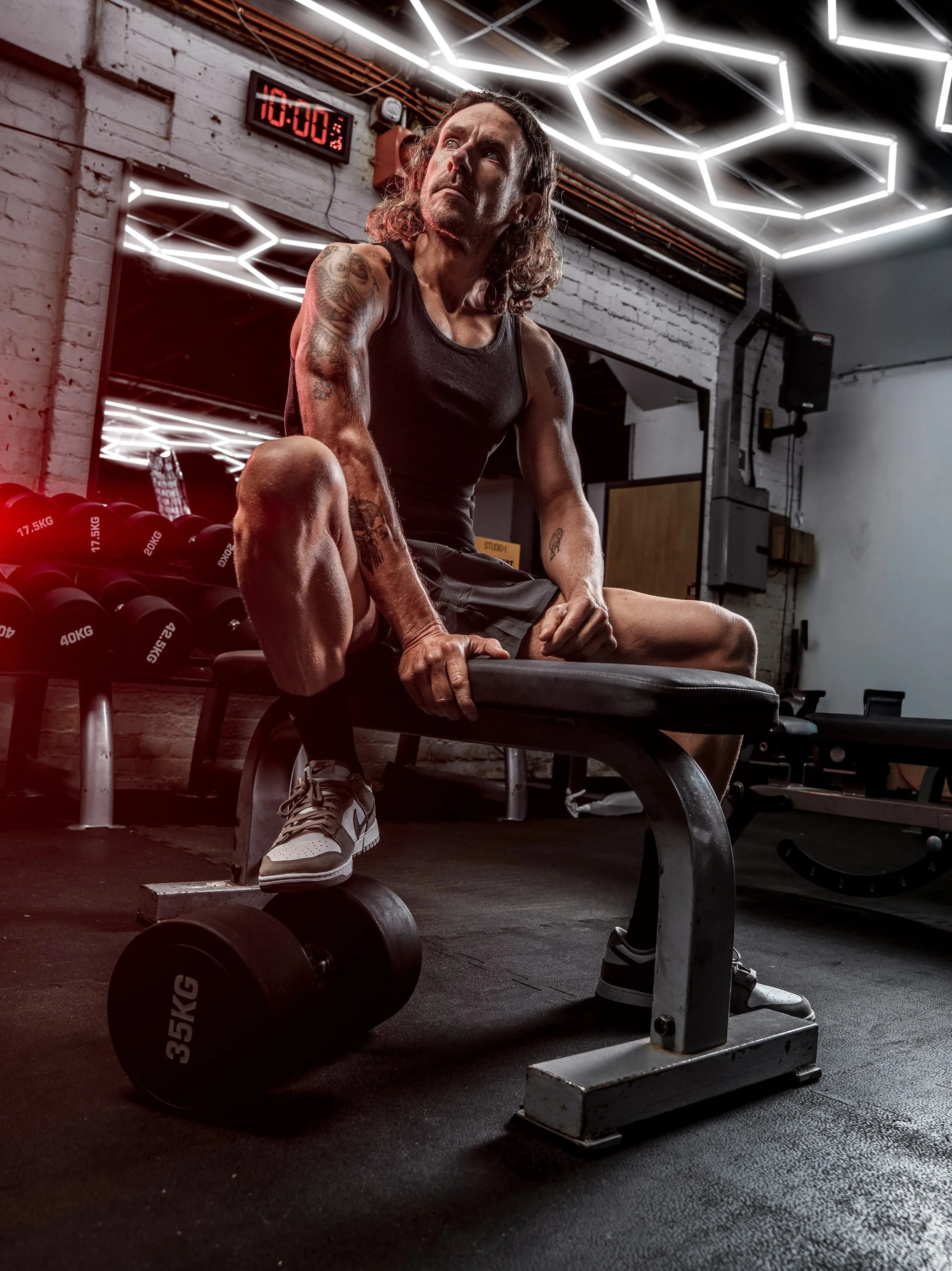 A muscular man with tattoos on his arms, wearing a black tank top and shorts, sitting on a bench in a gym, resting after a workout. The gym has exposed brick walls, a large mirror, and hexagonal LED ceiling lights. Dumbbells are visible in the backgr