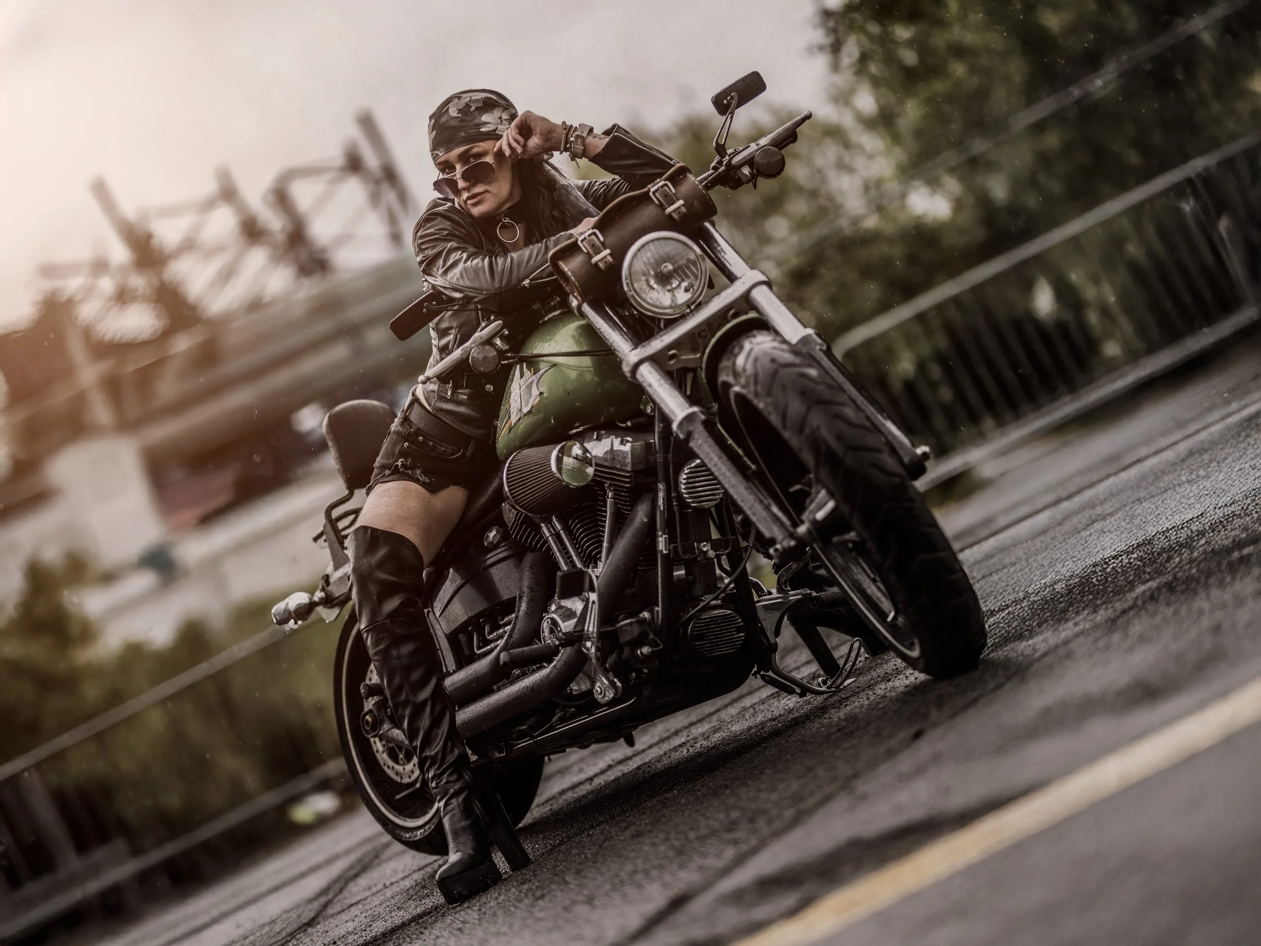 A woman wearing sunglasses, a leather jacket, and black shorts riding a black and green motorcycle on a street near a bridge and trees.