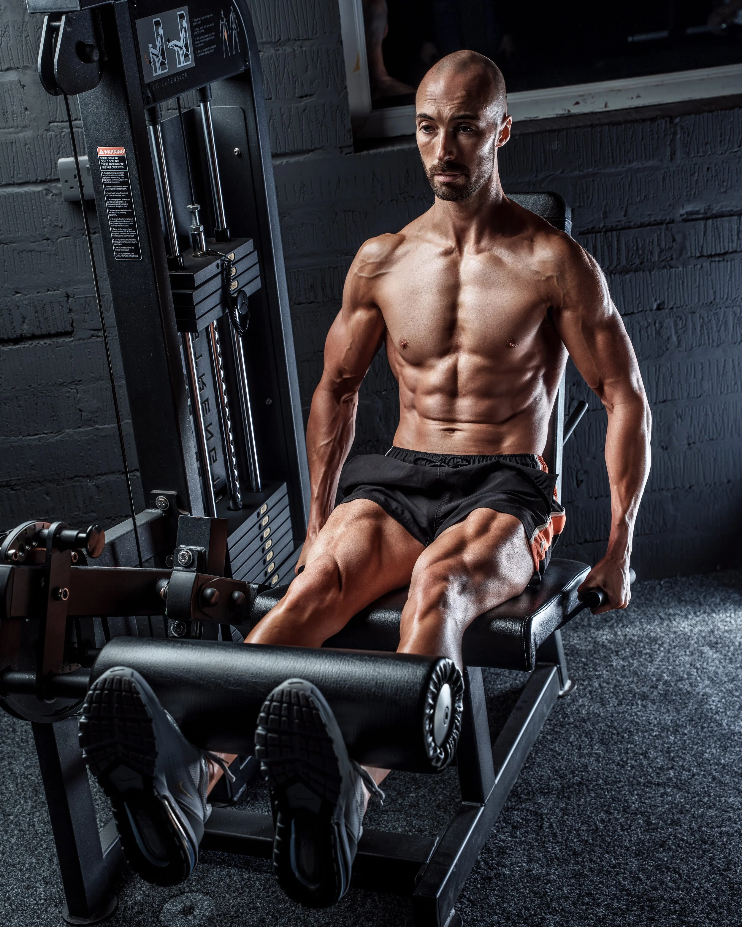 A muscular man with a shaved head and beard, shirtless, sitting on a leg curl machine in a gym, wearing black shorts and running shoes.