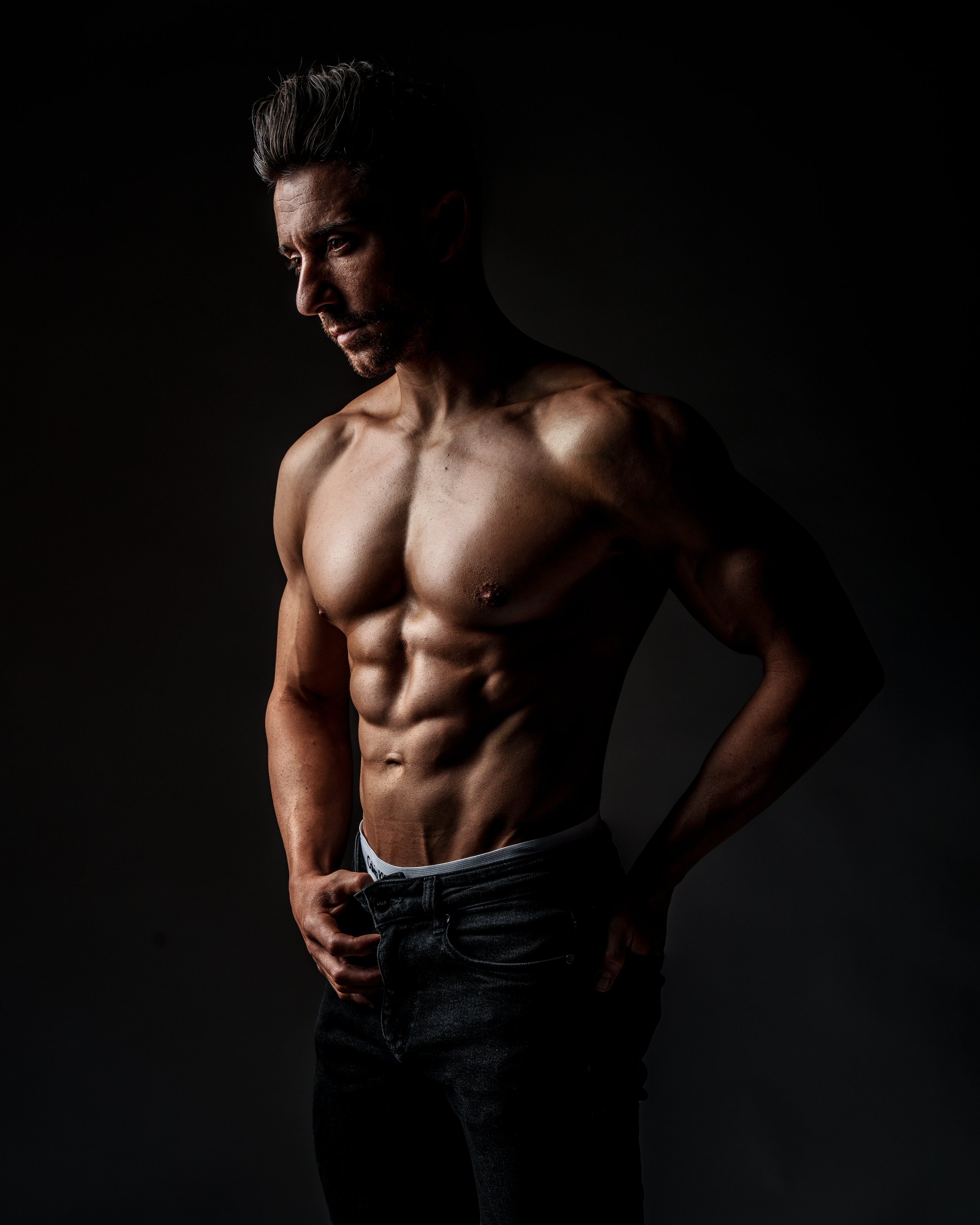 Low light fitness portrait of a shirtless muscular man posing against a dark background, highlighting physique definition.