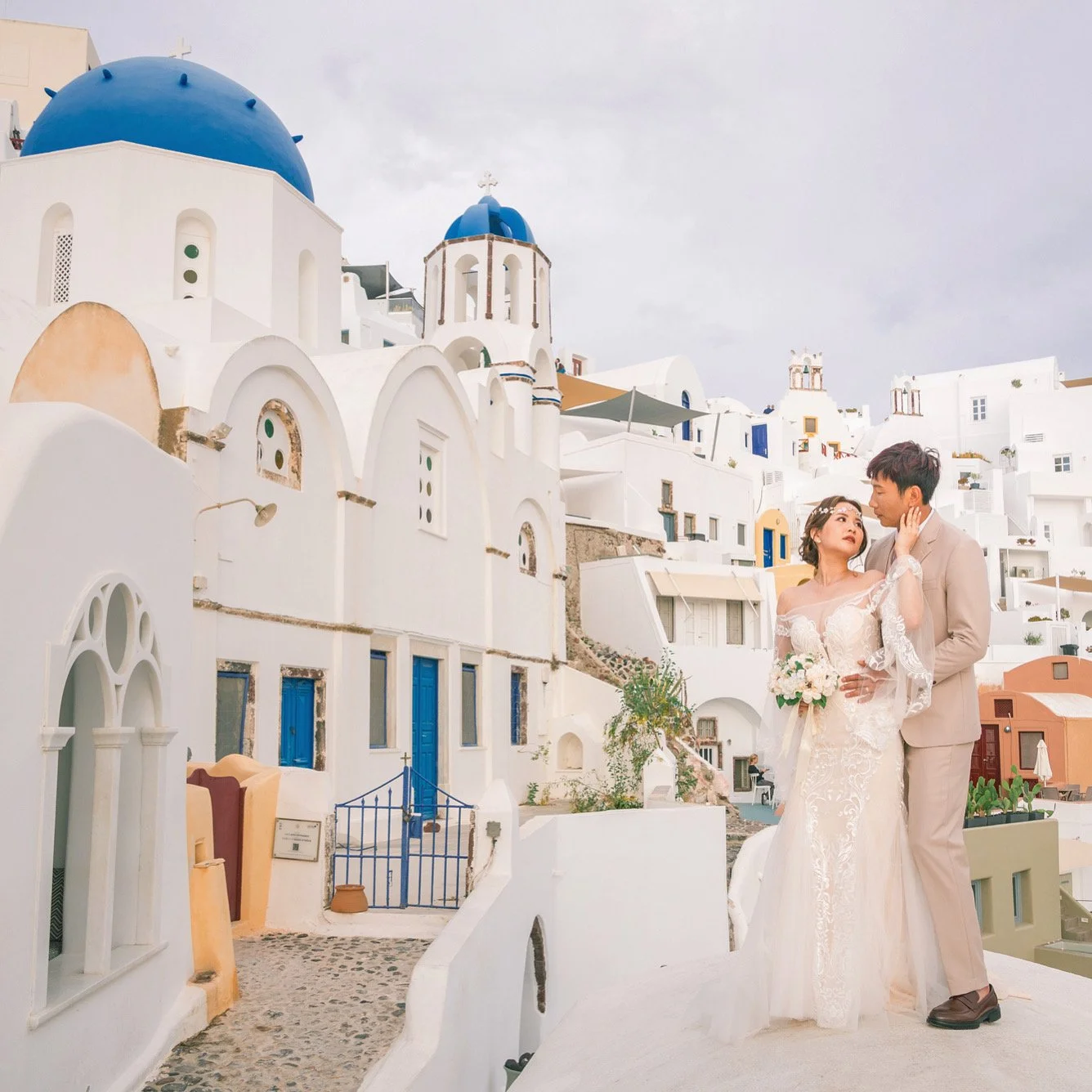 藍頂教堂旁的隱藏巷弄婚紗：白牆藍窗與花盆前景，私房 Santorini photoshoot 構圖；Hidden alley near blue domes wedding — white walls, blue shutters and potted foregrounds for private Santorini photoshoot compositions.