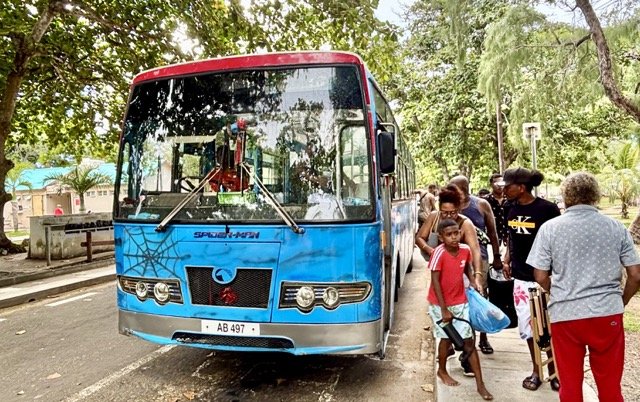 Port Louis Victoria Square 公車站熱鬧景象，旅客查詢路線並準備小額現金上車購票。 Bustling Port Louis Victoria Square bus stop with travelers checking routes and preparing small cash fares onboard.