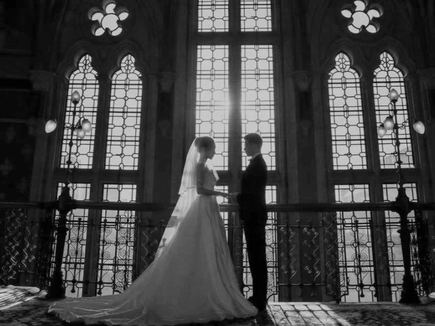Black and white photo of a bride and groom holding hands in front of large stained glass windows inside a grand building, probably a church or cathedral, during a wedding ceremony.