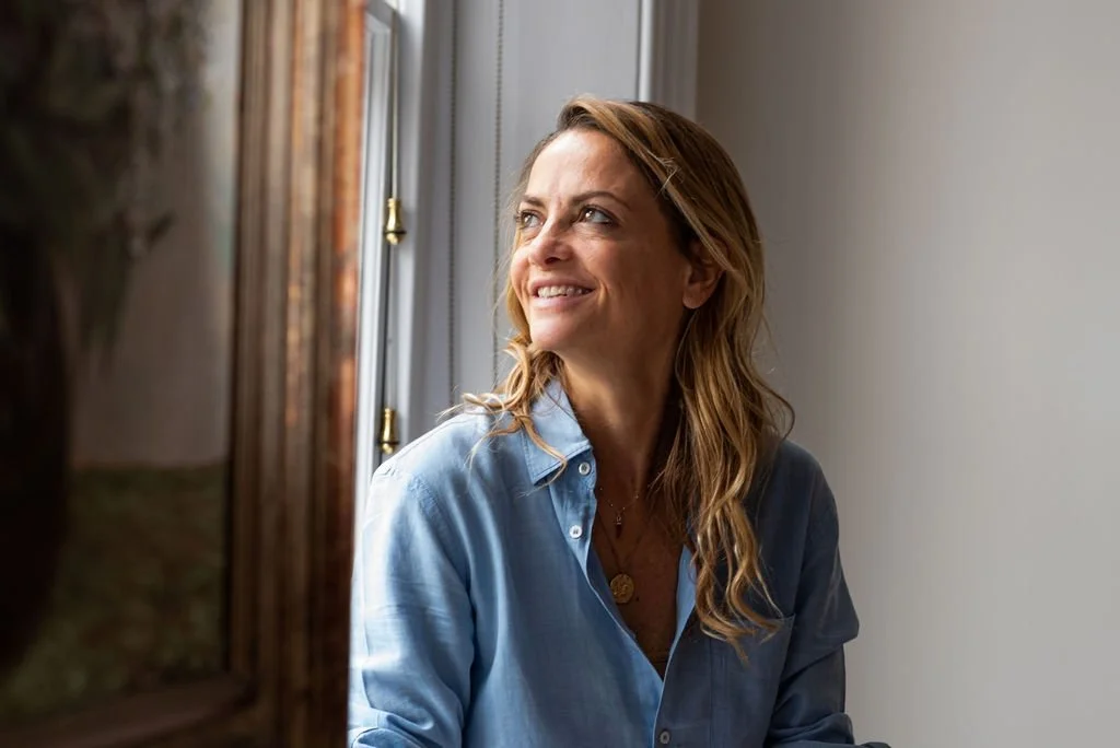 A woman with wavy blonde hair is smiling while looking out a window, wearing a light blue button-up shirt.