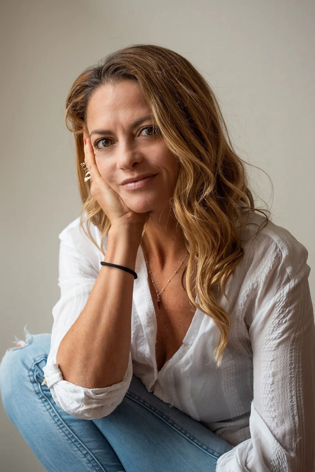 A woman with wavy, light brown hair and light skin is sitting and resting her head on her hand, slightly smiling at the camera. She is wearing a white blouse, blue jeans, and jewelry.
