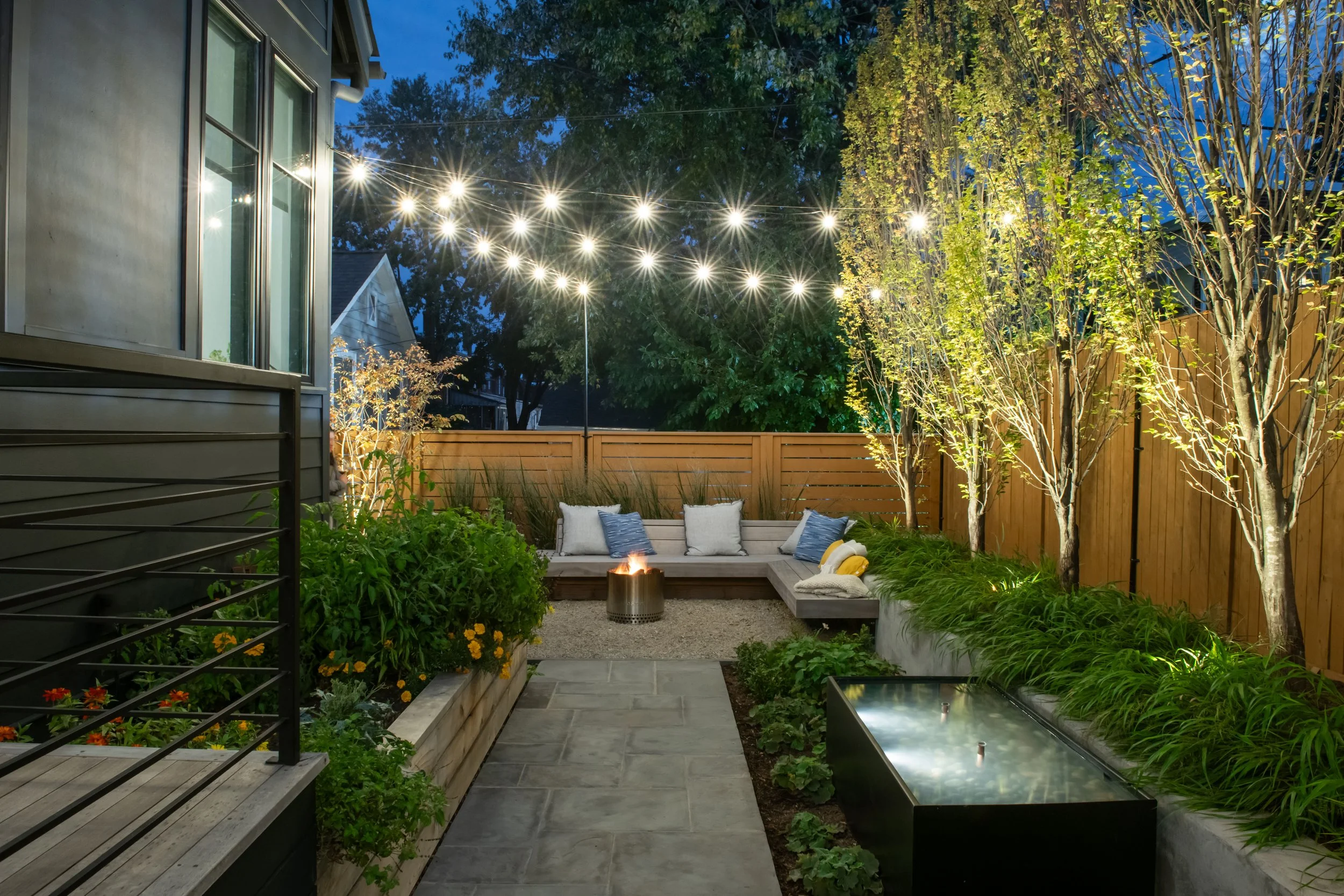 Bistro lighting over a custom bench in back yard of modern farmhouse provides illumination and contrast against this black modern farm house home in Alexandria, Virginia.  Designed by landscape architects, FORM Garden Design.  Custom fountain by DC P