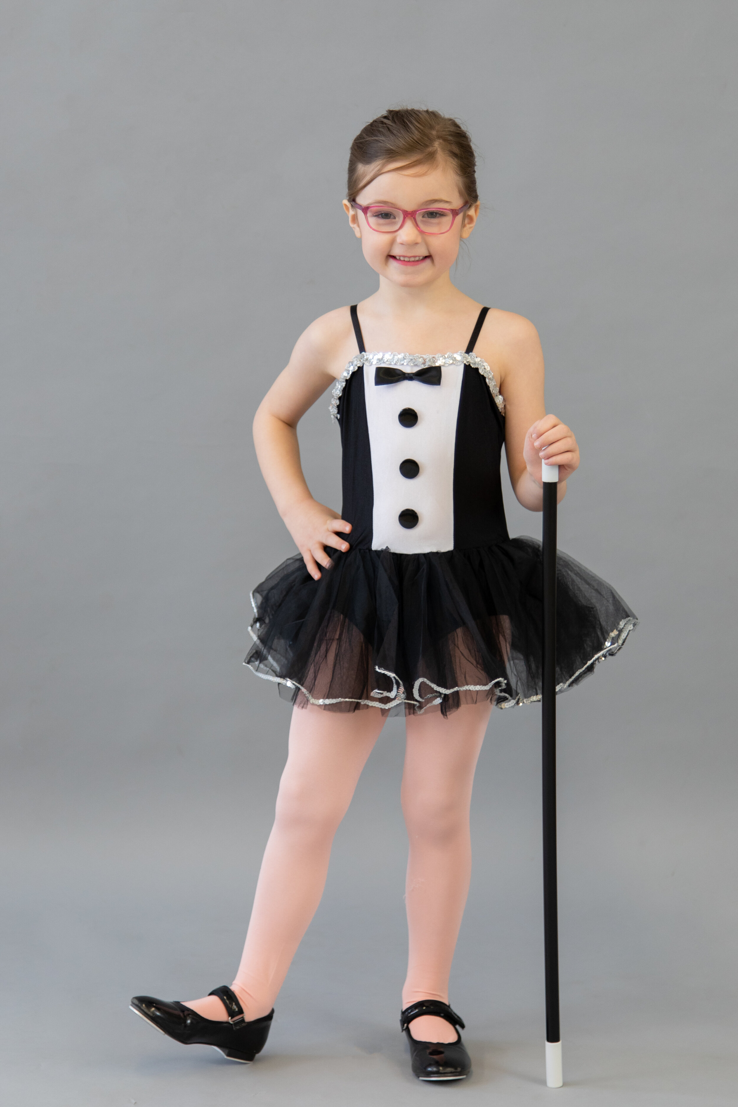 A young girl wearing a black and white tuxedo-themed tutu dress with a bow tie, pink glasses, black shoes, and holding a white cane, standing against a plain gray background.
