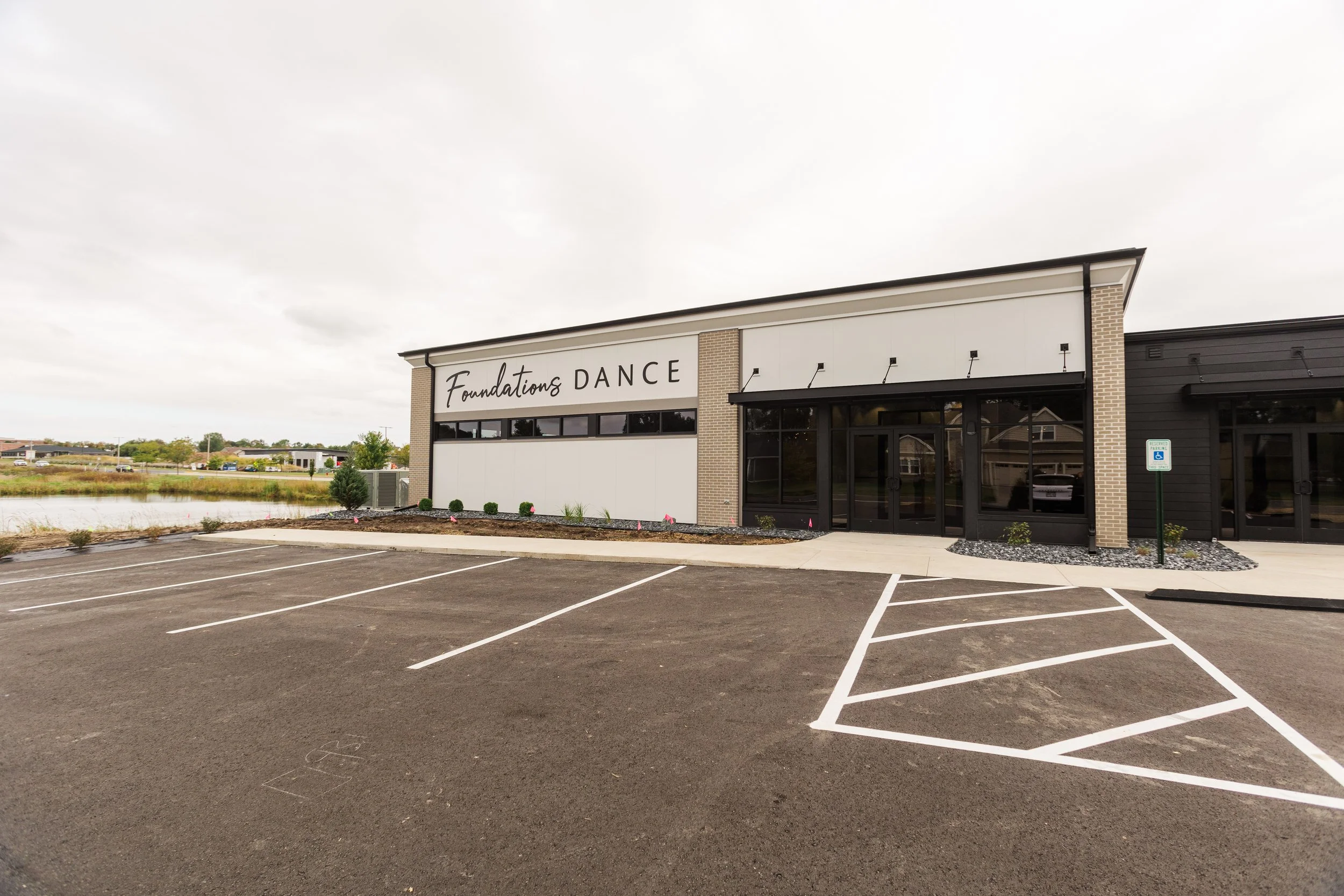 Exterior view of a dance studio called Foundations Dance, with an empty parking lot and a cloudy sky.