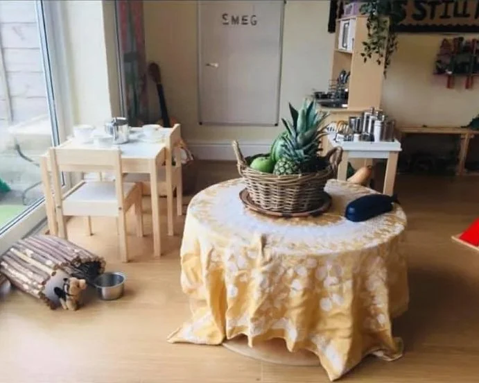 A round table covered with a yellow tablecloth, holding a basket of pineapples and other fruits, in a bright room with a small dining set and a door with the word 'SMEG' on it.