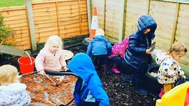 Children playing in a sandbox and exploring in an outdoor play area with wooden fencing, a traffic cone, and various toys.