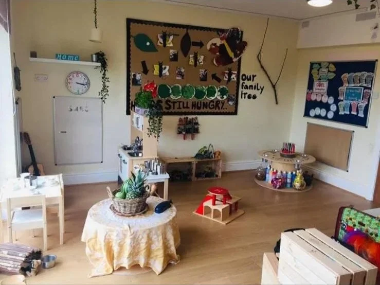 A cozy indoor kids' playroom with educational decorations, a table with a basket of plastic fruits, small furniture, and wall art including a bulletin board and a small blackboard.